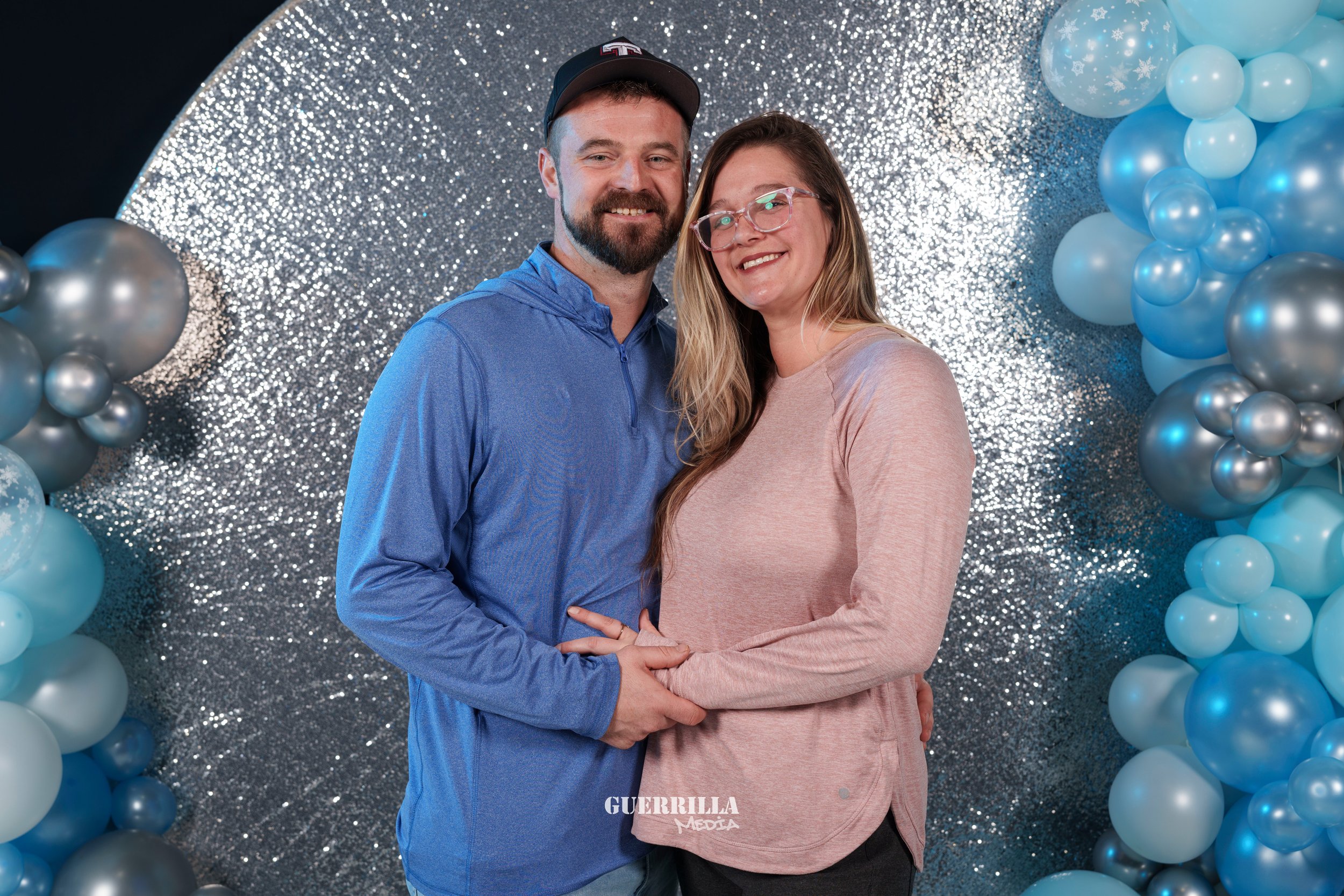 A smiling couple standing close together in front of a sparkling silver backdrop and blue balloons, with the man wearing a blue shirt and a baseball cap, and the woman wearing glasses and a pink top.