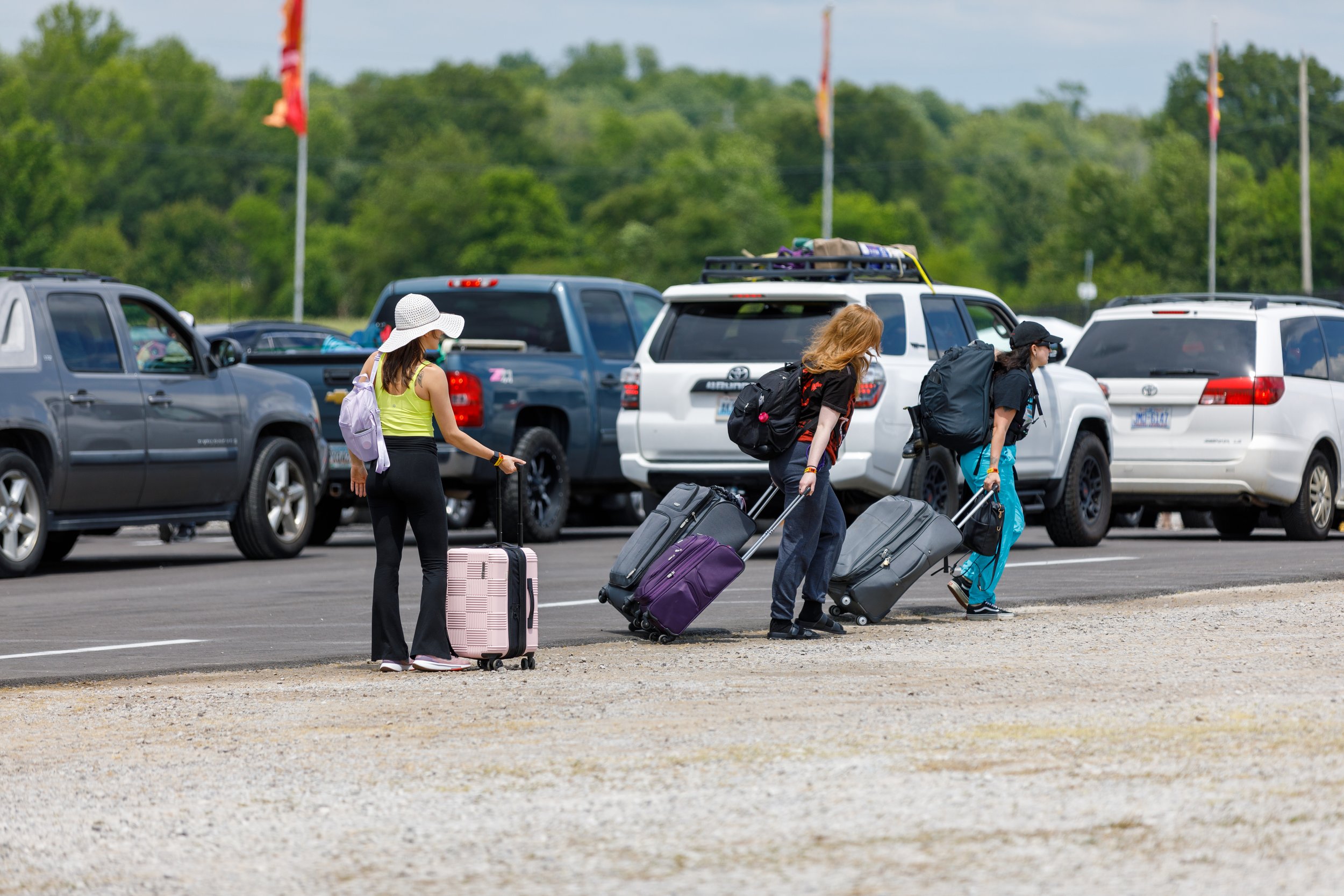 Three women walk with rolling suitcases in a parking lot, with parked cars and green trees in the background on a partly cloudy day.