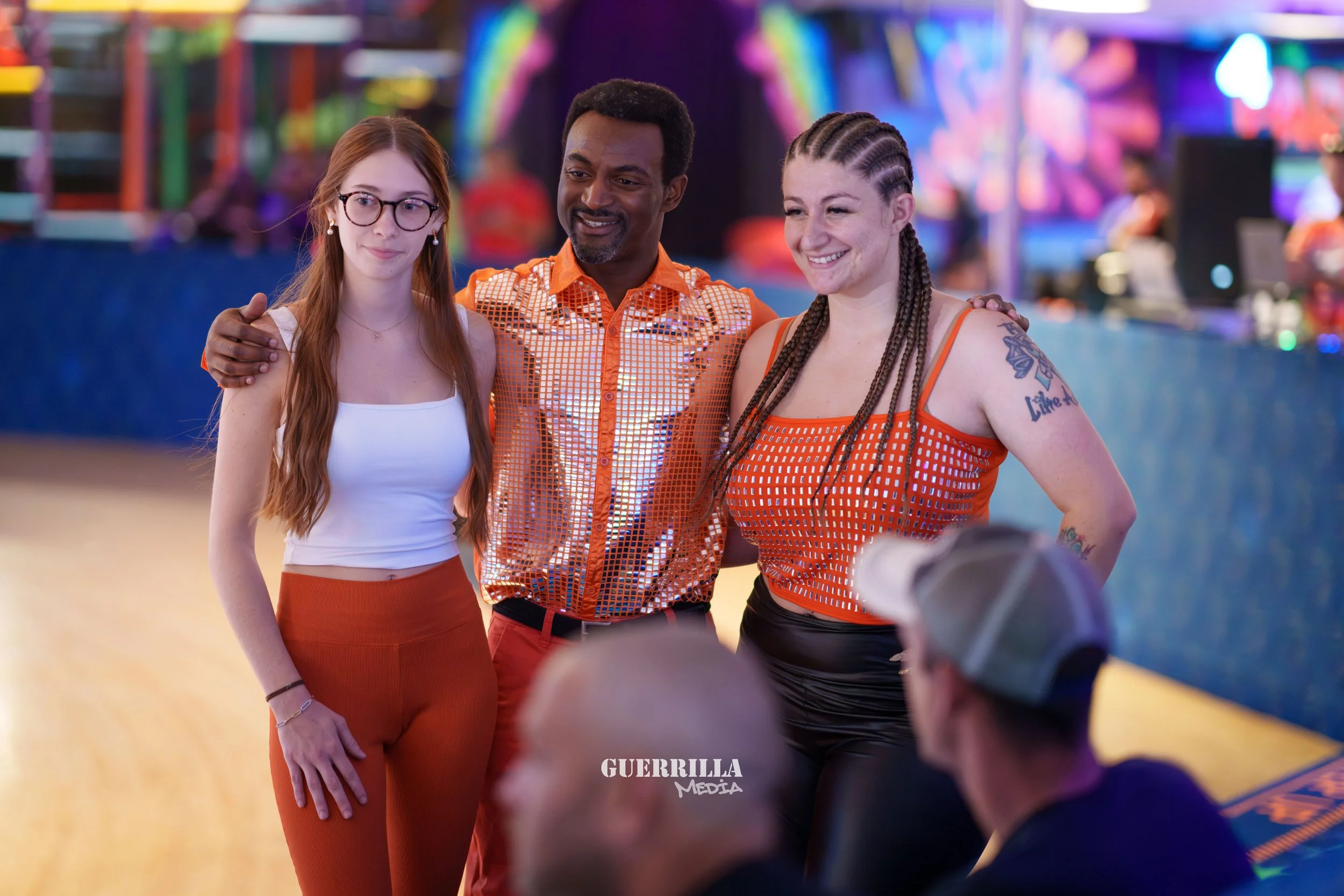 Three people posing for a photo at a roller skating rink, with colorful lights and a bar in the background.