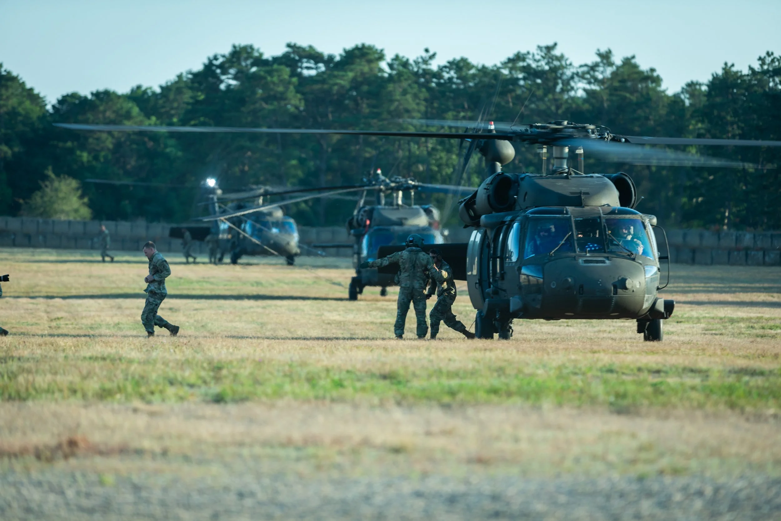 Military personnel preparing helicopters on a tarmac with trees in the background.
