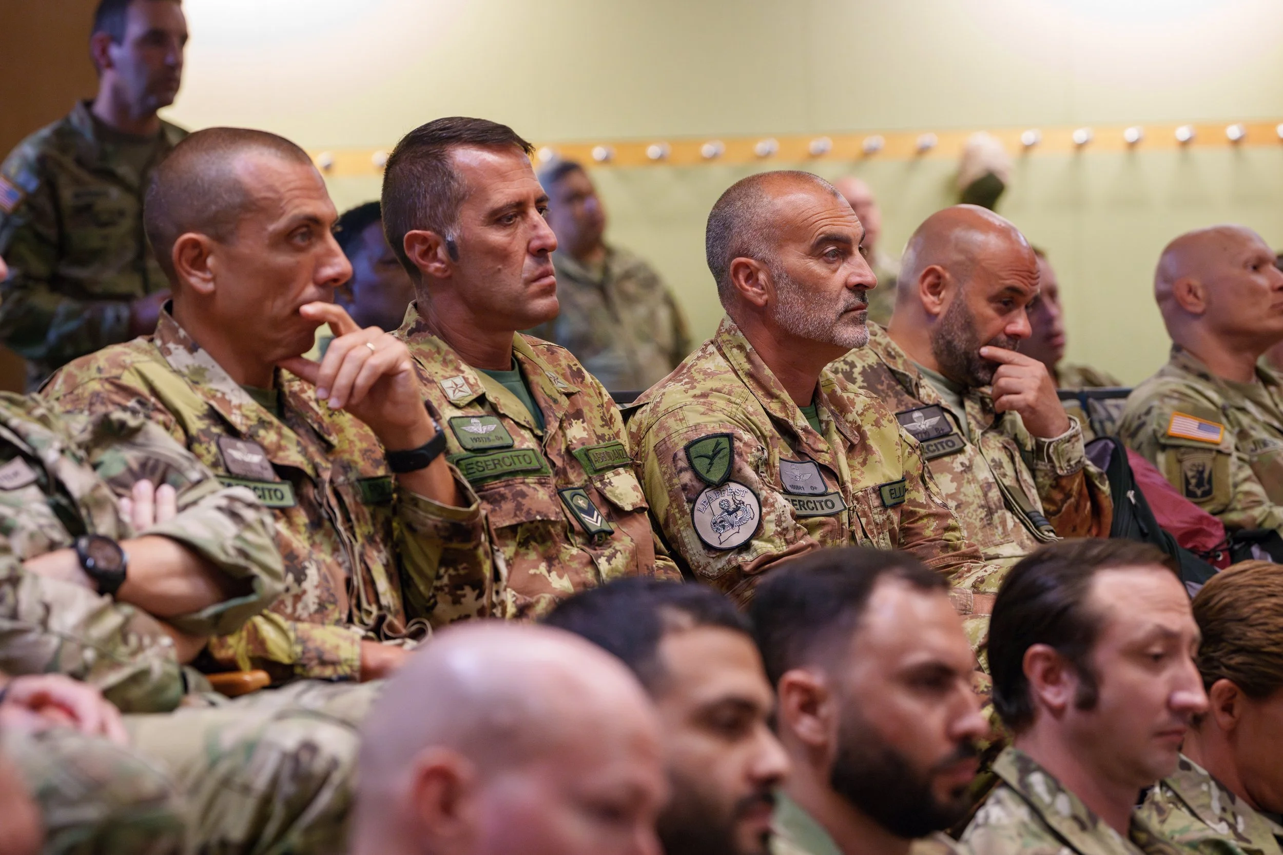 Group of military personnel attending a meeting or briefing, wearing camouflage uniforms, in a room with yellowish walls.