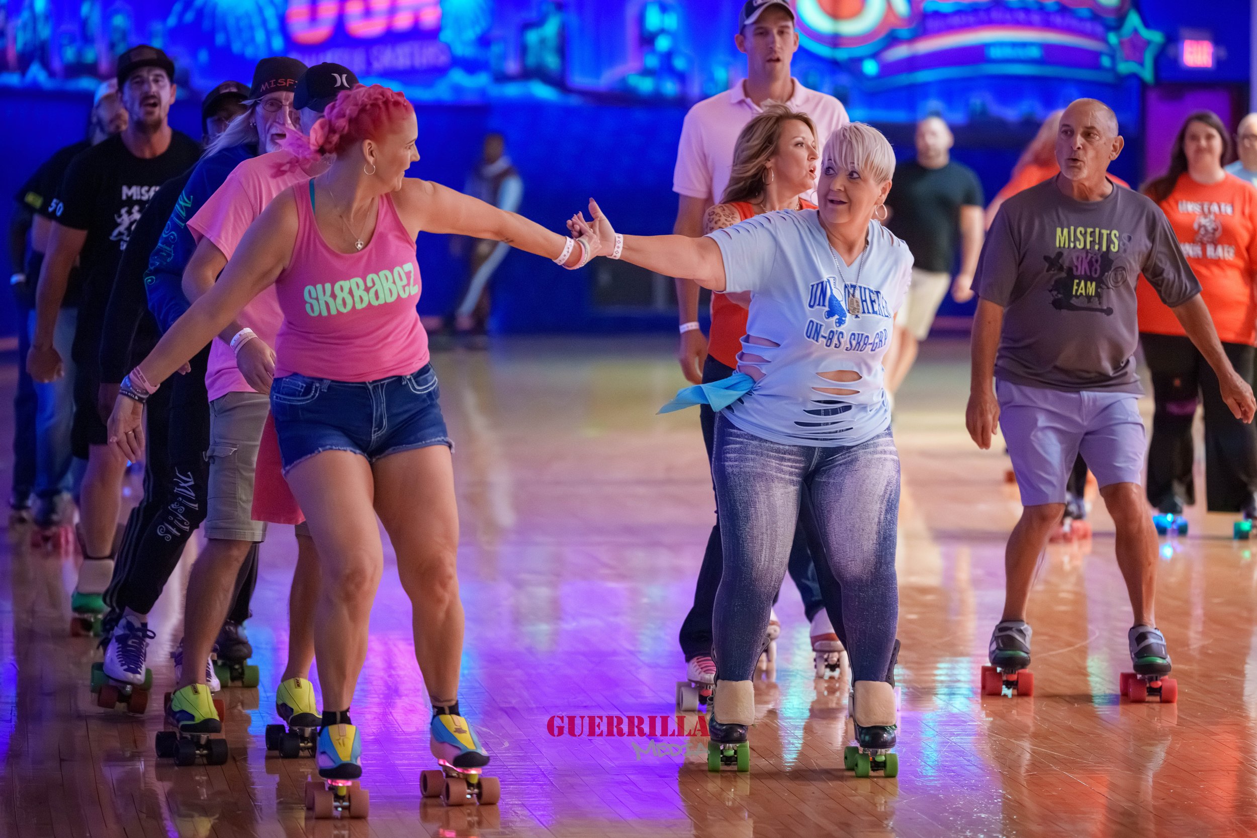 Group of people roller skating indoors, some stop and support each other, with colorful neon lights in the background.
