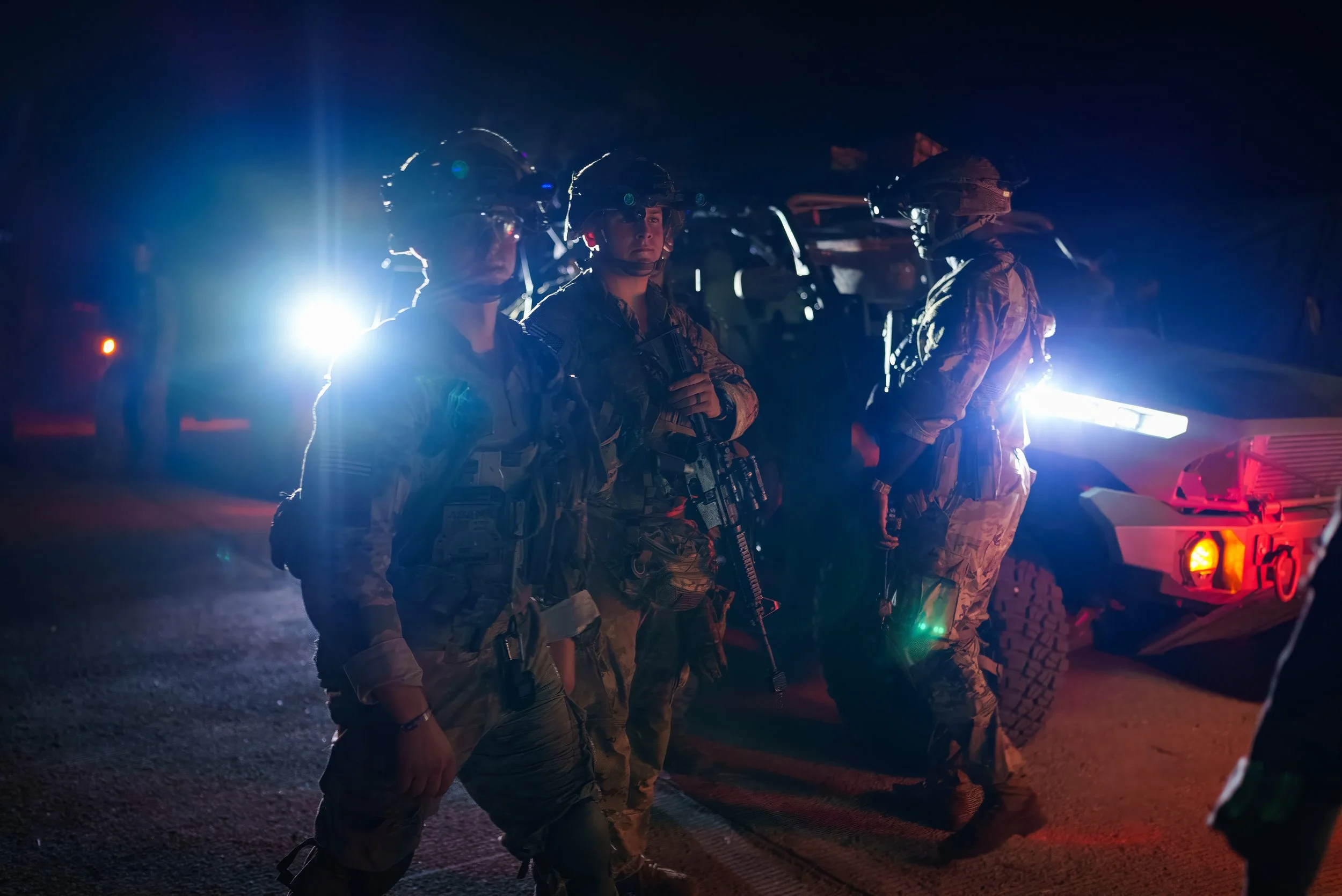 Group of soldiers in combat gear standing in front of military vehicles at night with bright lights shining behind them.