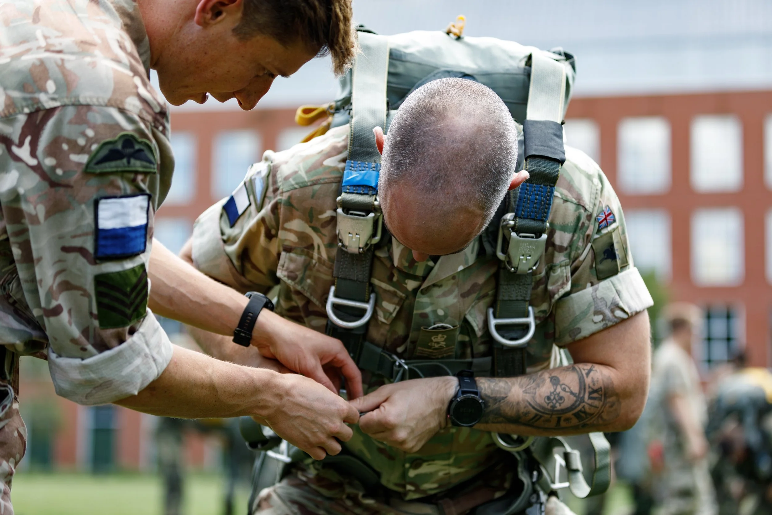 Two soldiers in camouflage uniforms are fastening a piece of equipment while standing on a grassy field, with other soldiers and a brick building in the background.