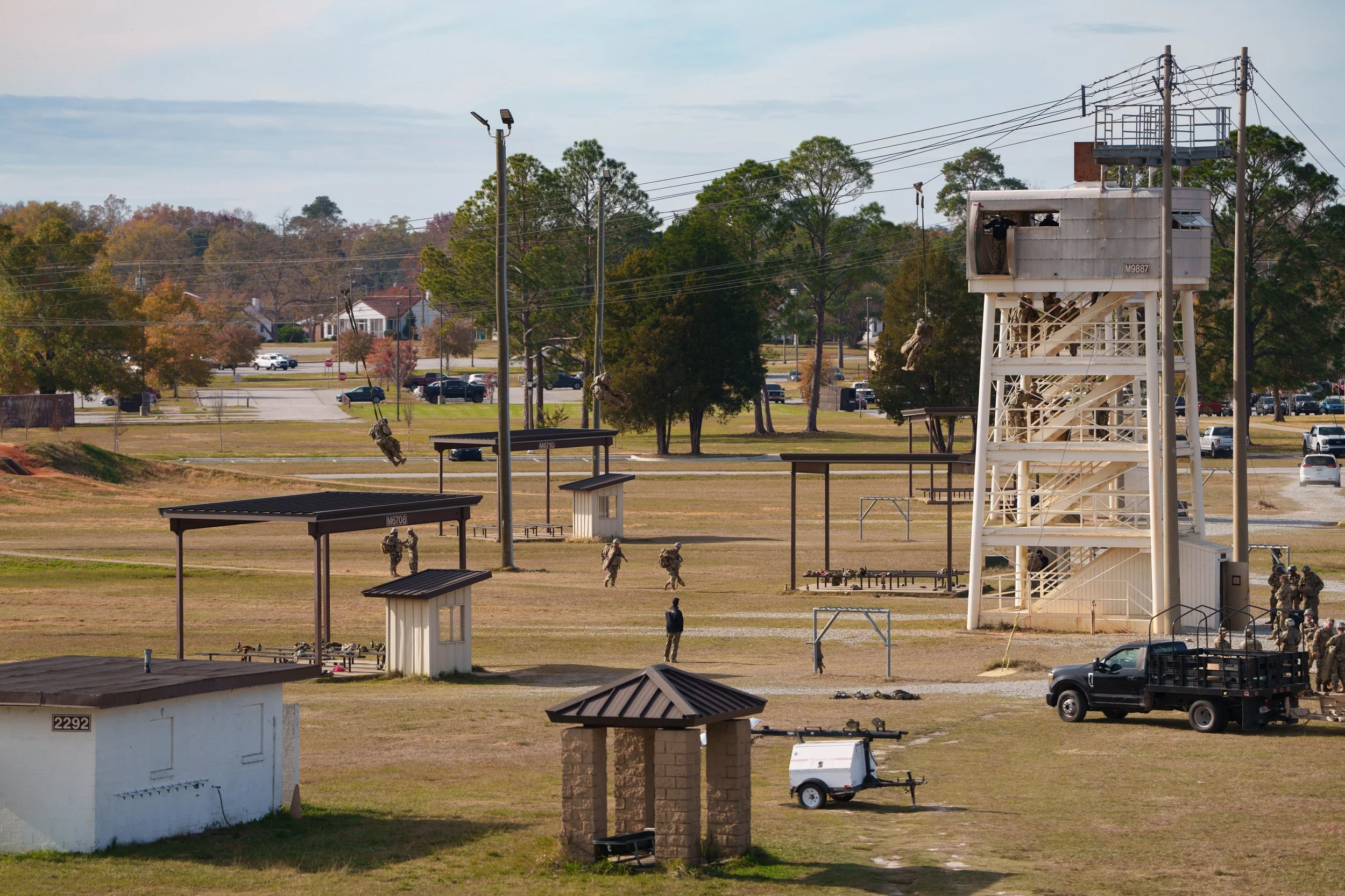 Military soldiers practicing in an outdoor training area with a tall white metal observation tower, covered benches, a small shelter, and electrical poles. Trees and a parking lot are visible in the background.
