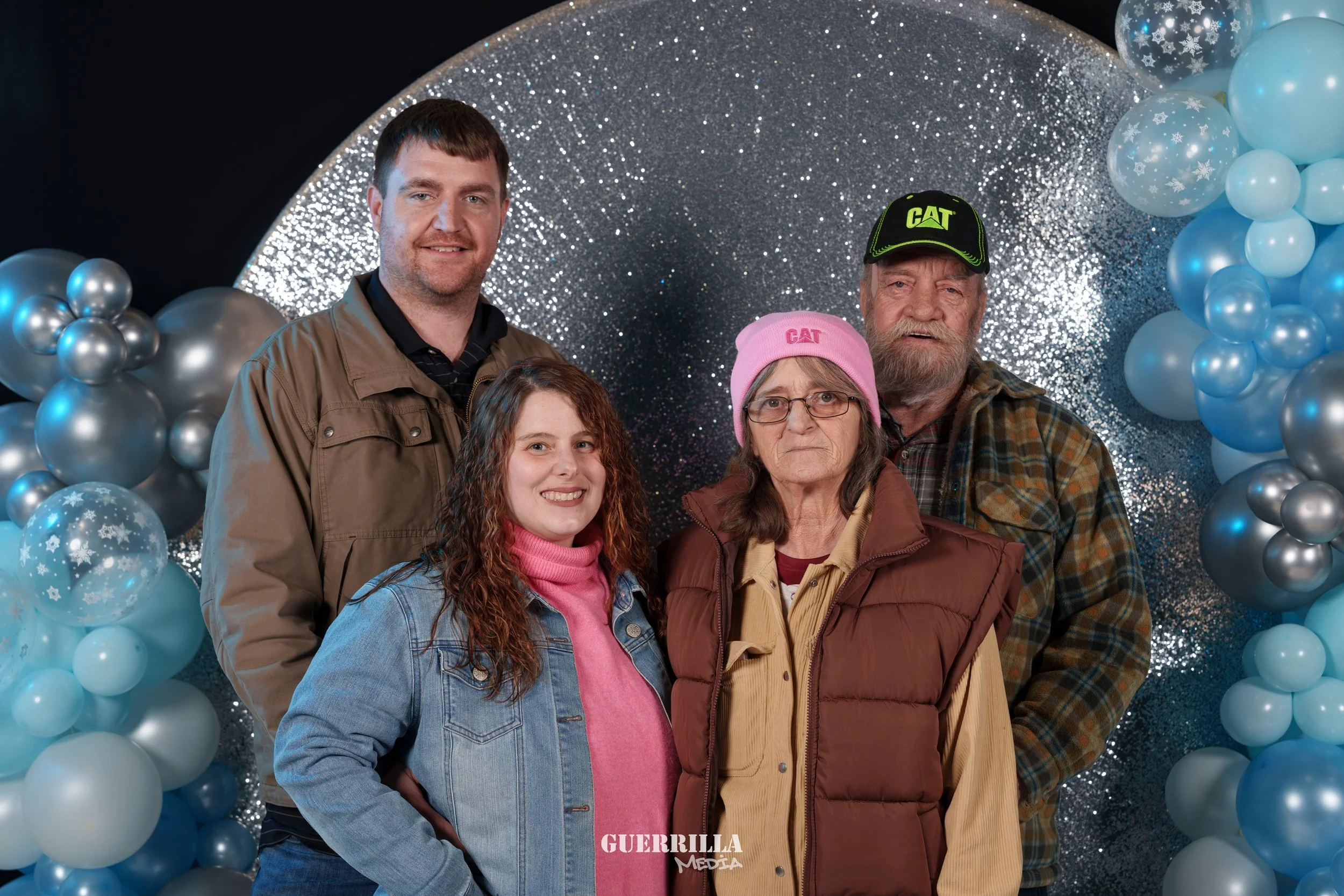 Group of four people posing in front of a silver and blue balloon backdrop, with one woman wearing a pink hat and a brown vest, a man wearing a black baseball cap and plaid shirt, a young woman in a pink turtleneck and denim jacket, and a man in a ta