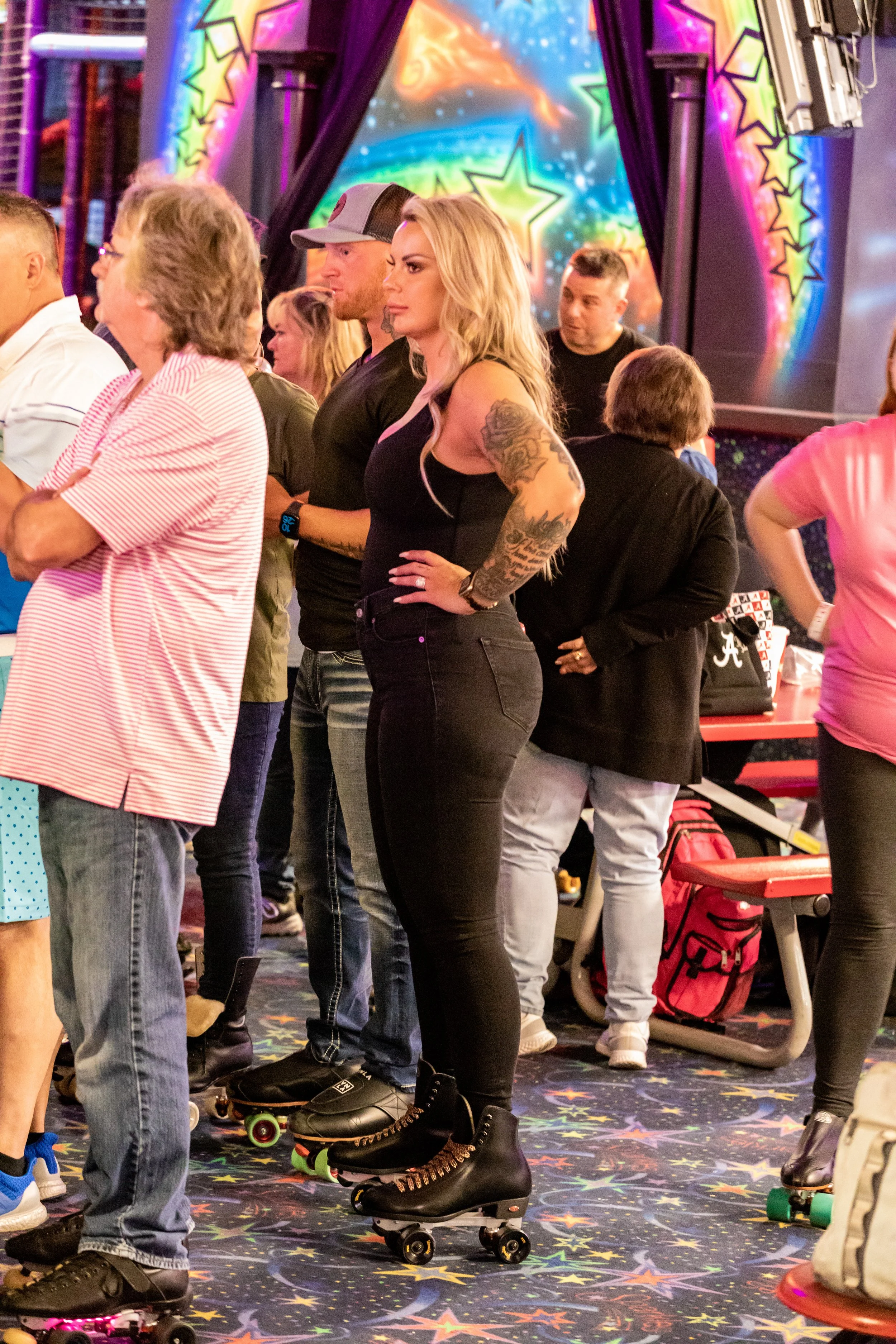 A group of people at a roller skating rink, with a colorful star-themed backdrop. The woman in the center has long blonde hair, tattoos on her arms, and black skating shoes. She is wearing a black top and black pants, with one hand on her hip.