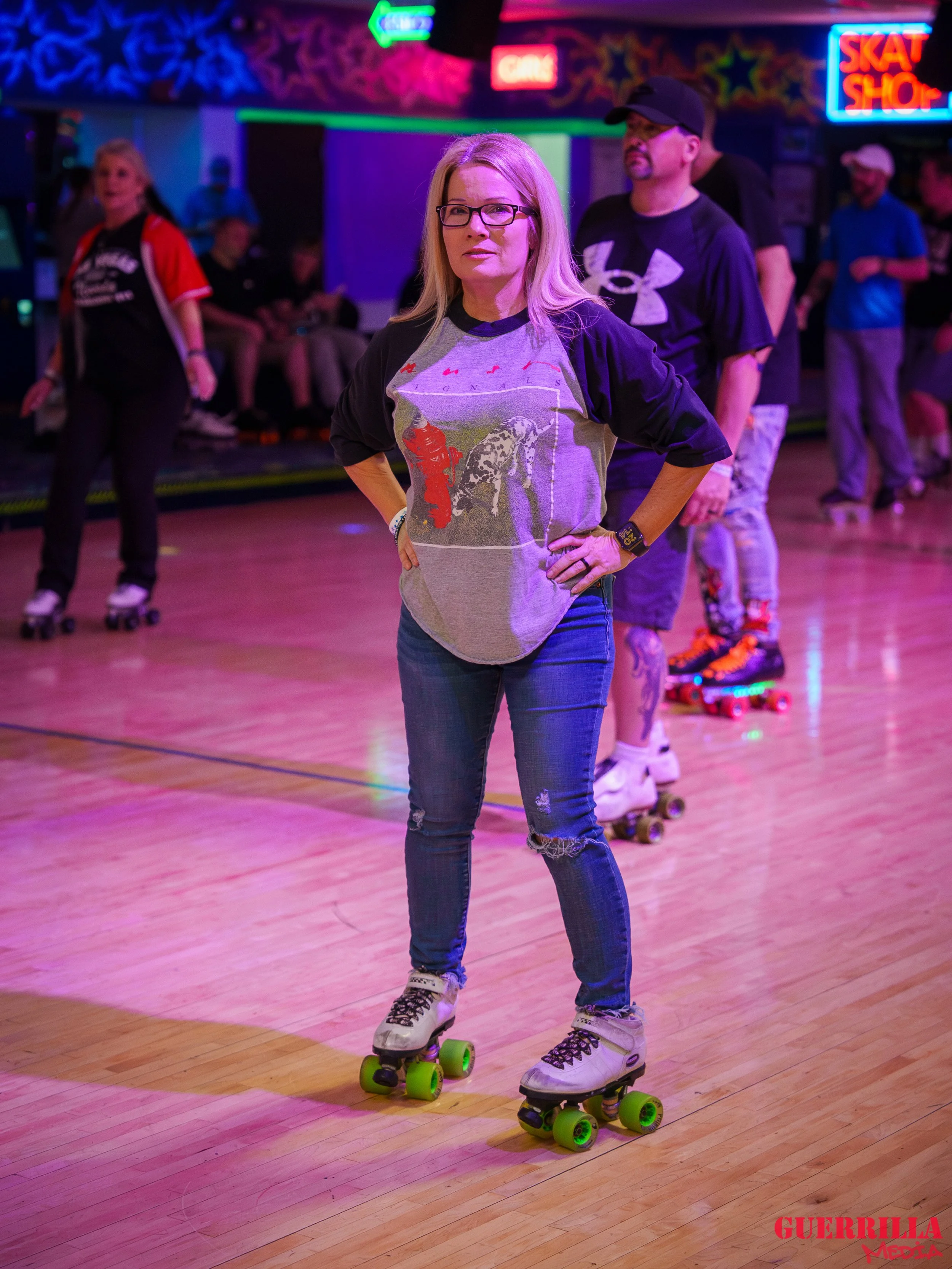 A woman with blonde hair and glasses standing on roller skates on a rink, with her hands on her hips, surrounded by people skating and neon signs in the background.