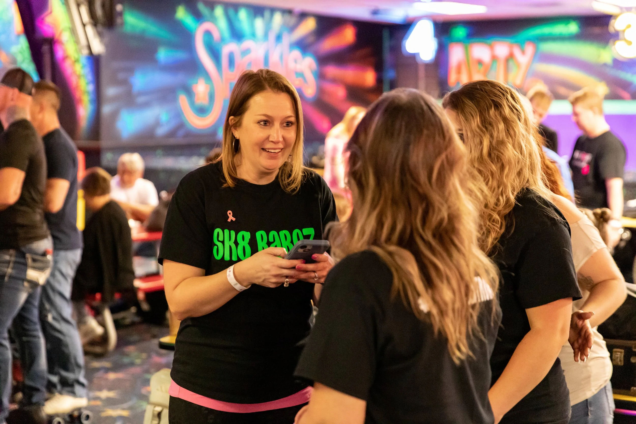 A group of women engaged in conversation at an arcade, with a colorful Neon sign in the background reading 'Sparkles' and 'Arena'.