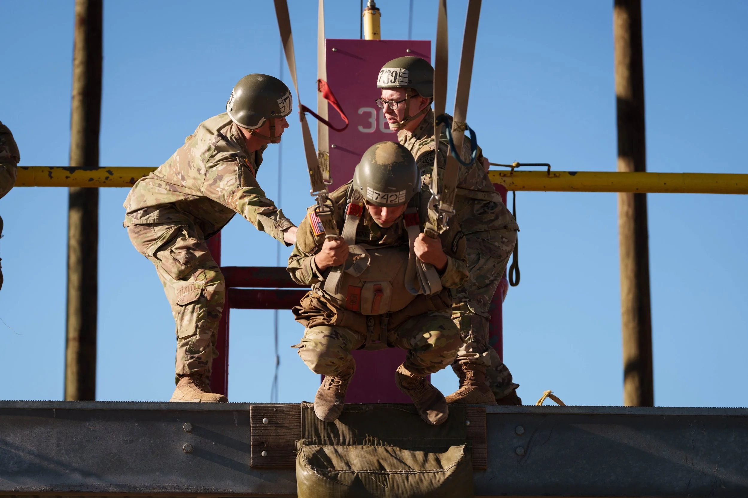 Three soldiers in military uniform and helmets assisting a fellow soldier to jump from a raised platform for a parachuting or fall training exercise, with clear blue sky in the background.