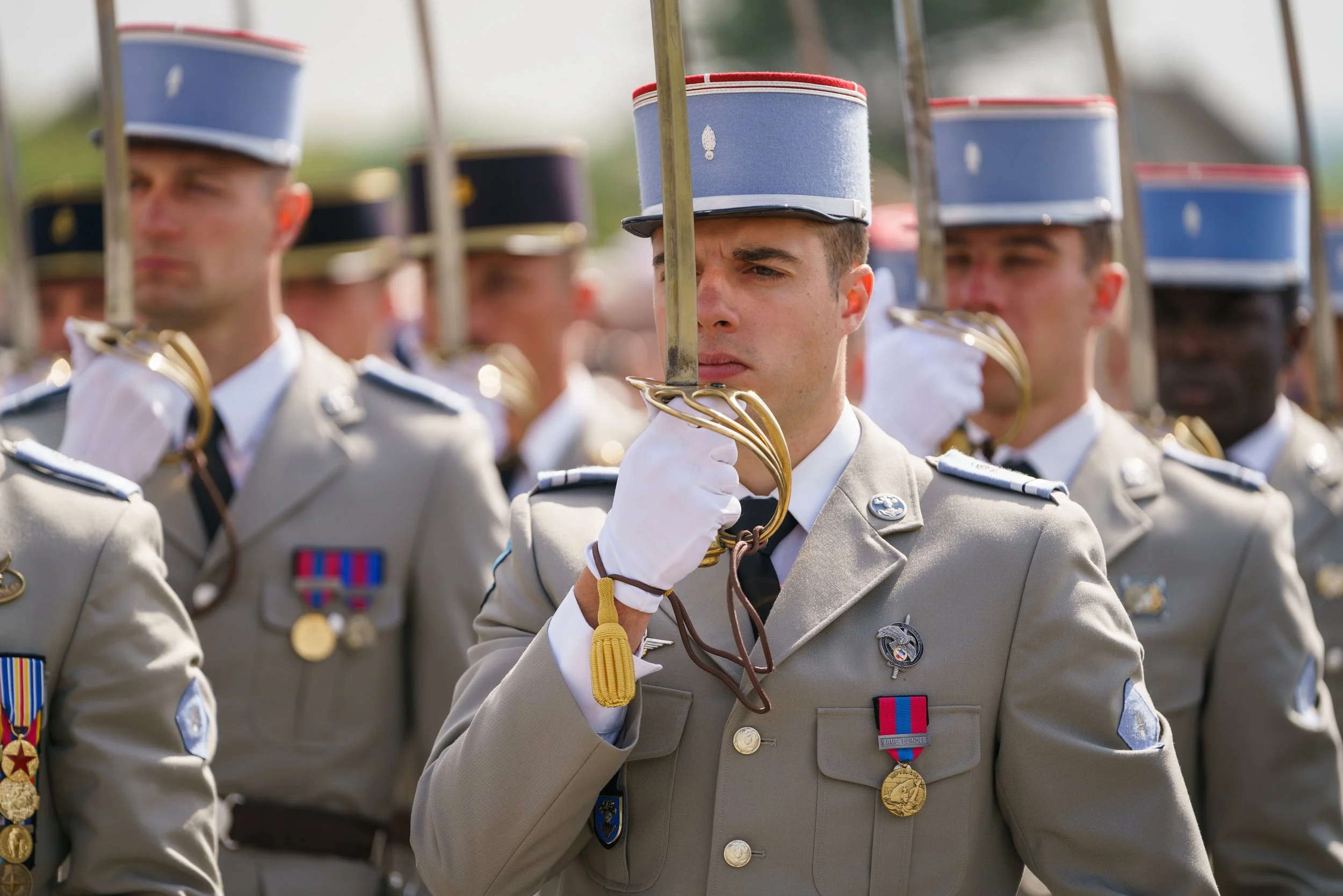 Group of military personnel in uniform during a ceremony, with a soldier in the front holding a ceremonial sword perpendicular to the ground, wearing medals and a hat with a blue band.