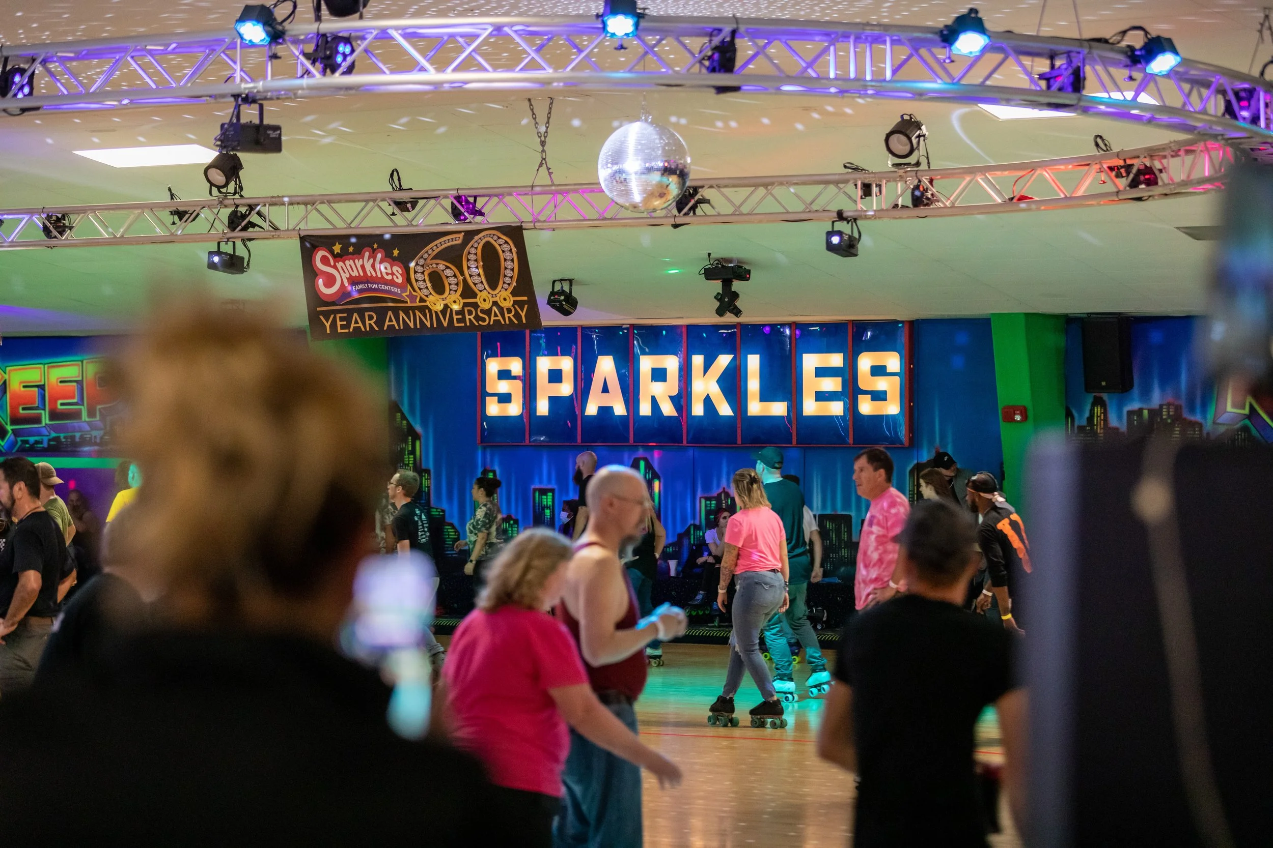 People roller skating at an indoor roller rink decorated for a 60th anniversary celebration with a sign that says 'SPARKLES' and a disco ball overhead.
