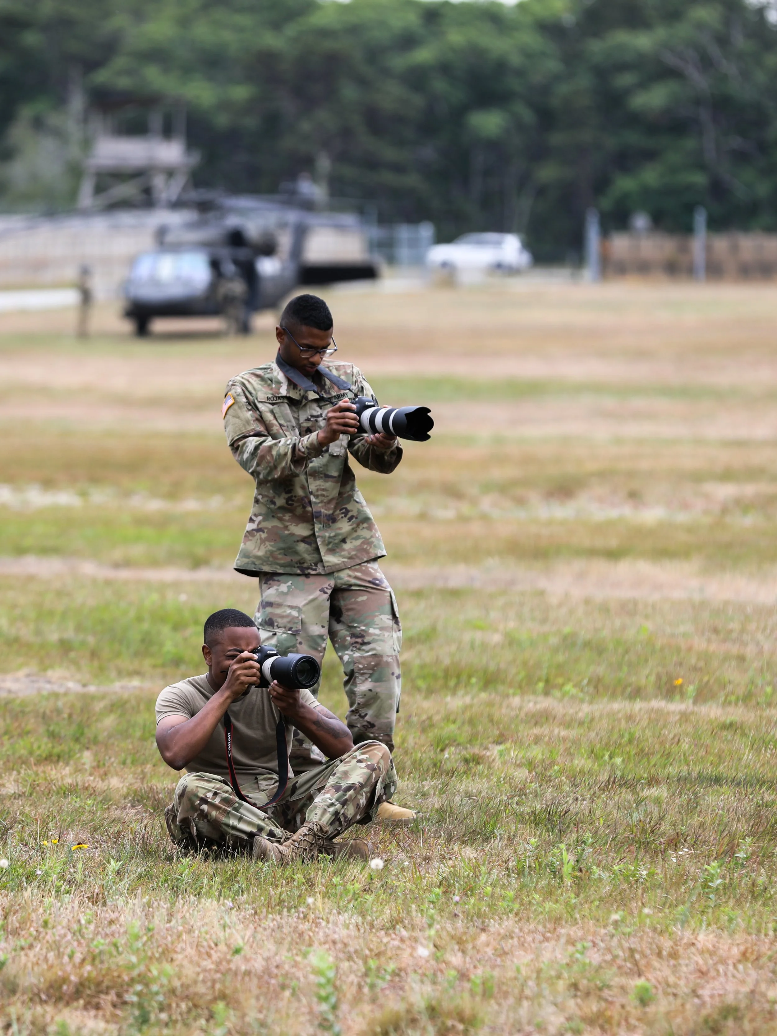 Two soldiers in camouflage uniforms taking photos with professional cameras in an open grassy field.