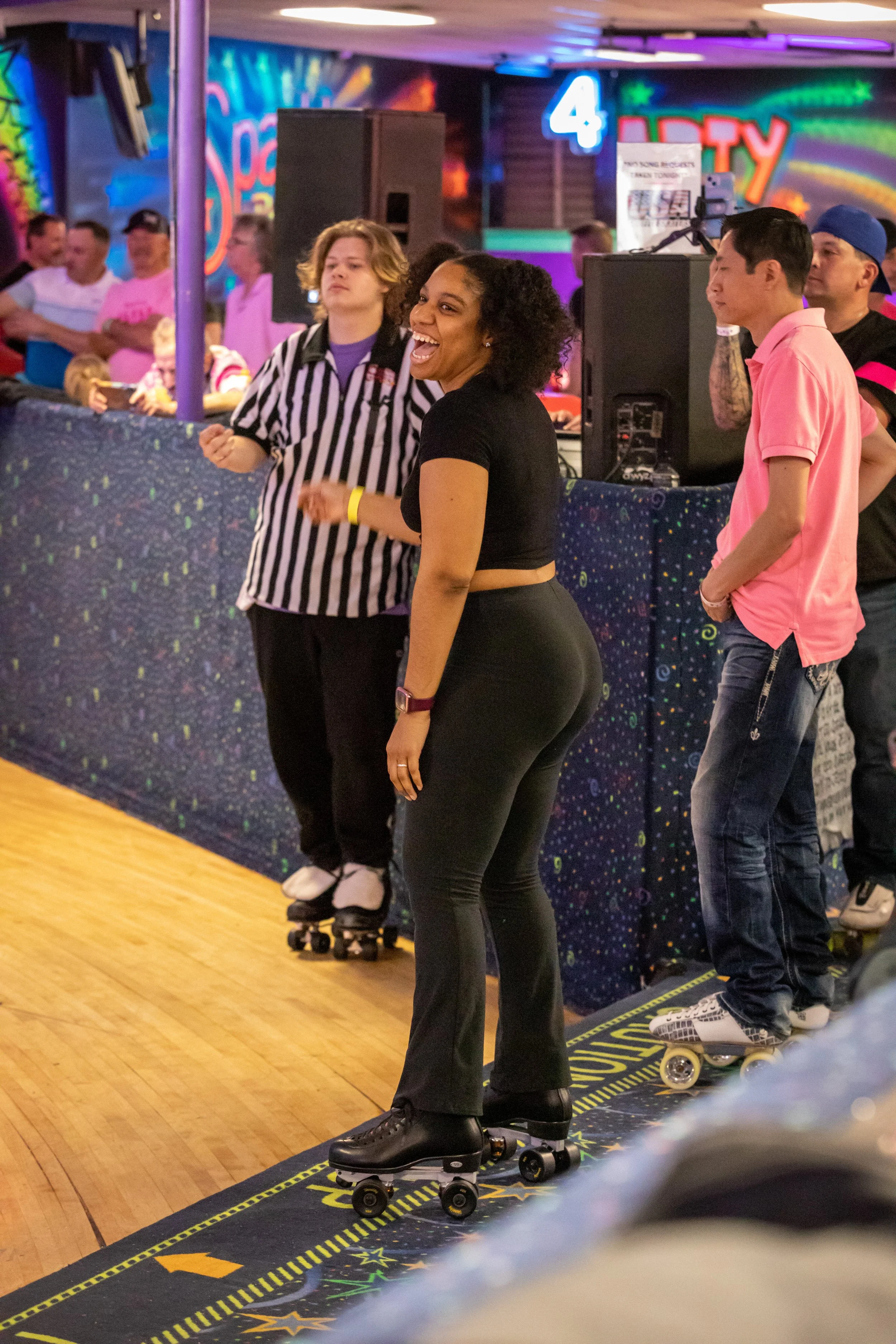 A woman laughing and riding roller skates at a fun indoor skating rink with colorful neon signs and a crowd of people in the background.