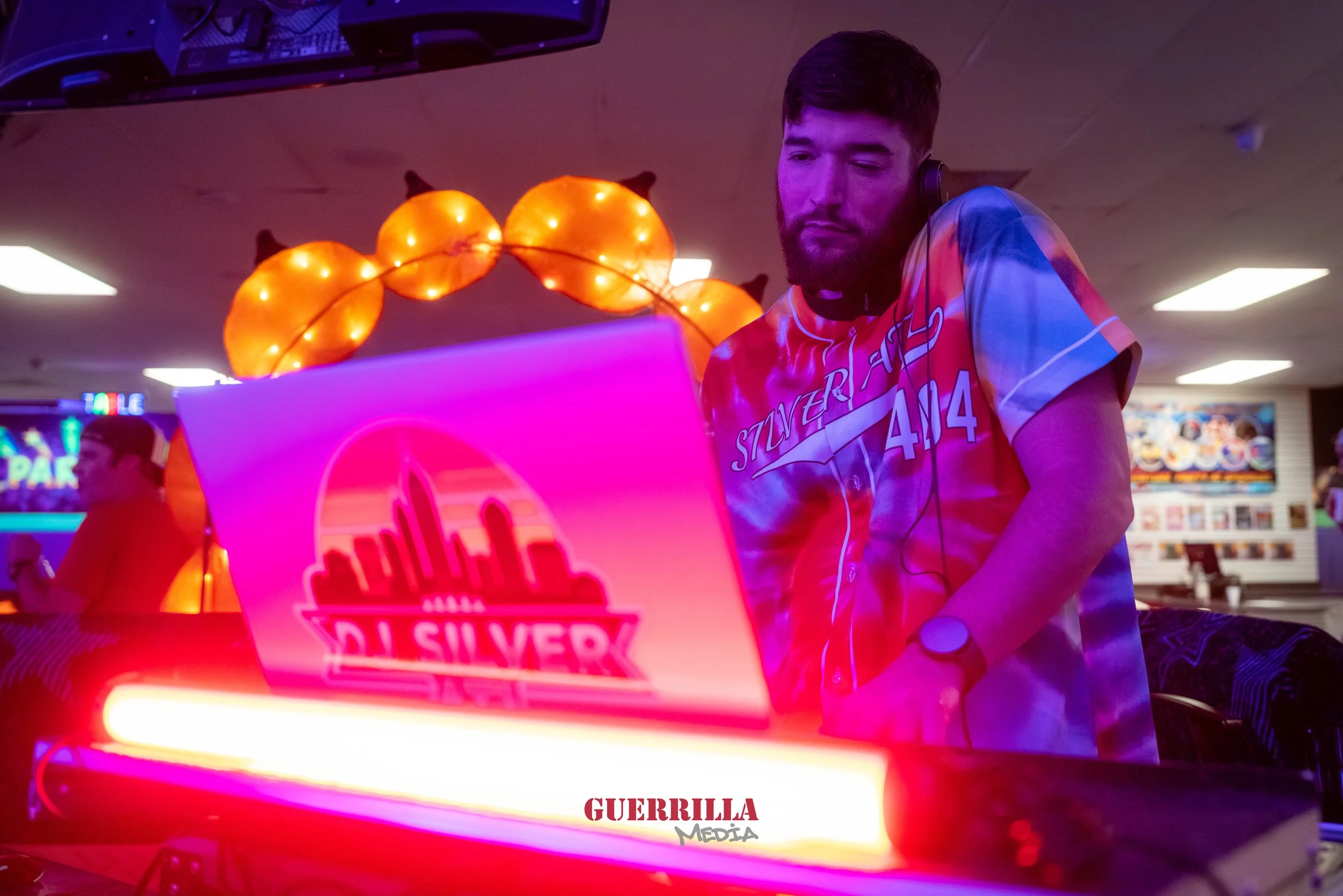 A young man with a beard wearing a colorful tie-dye shirt and a watch, DJing at an event with vibrant neon pink and orange lighting, a laptop with a cityscape logo, and decorative orange lamp or balloon sculpture in the background.