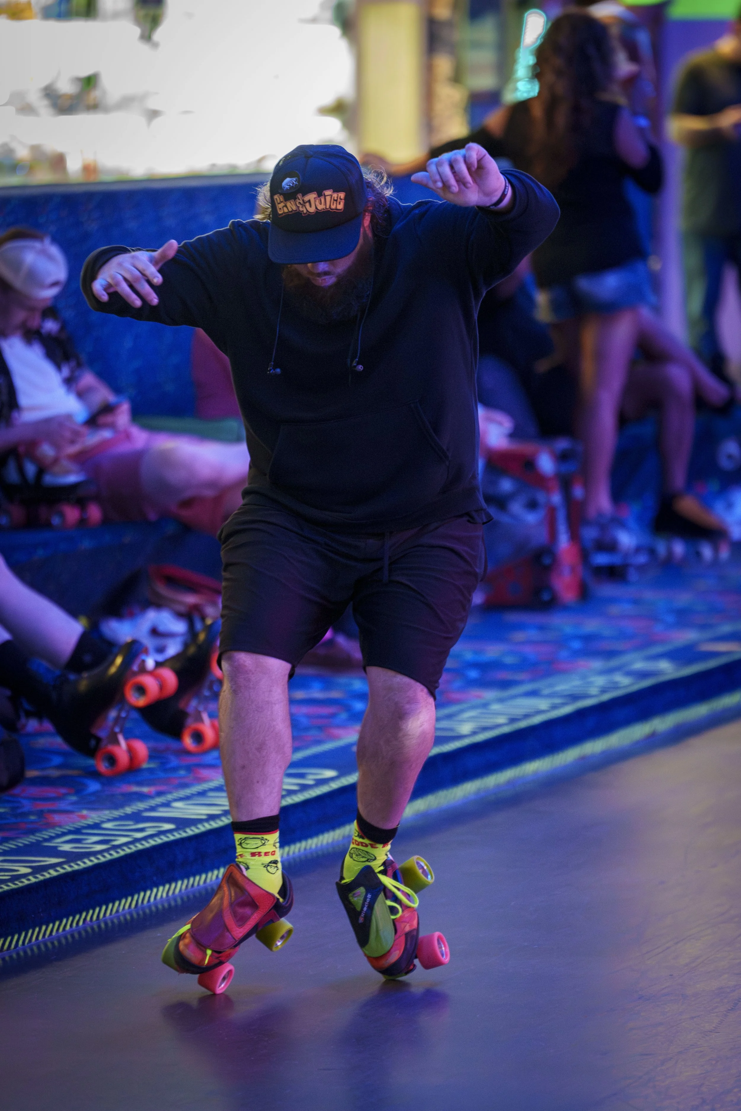 A man roller skating on a roller skating rink with colorful lighting, wearing a black hoodie, black shorts, a black cap, and yellow socks with skate shoes.