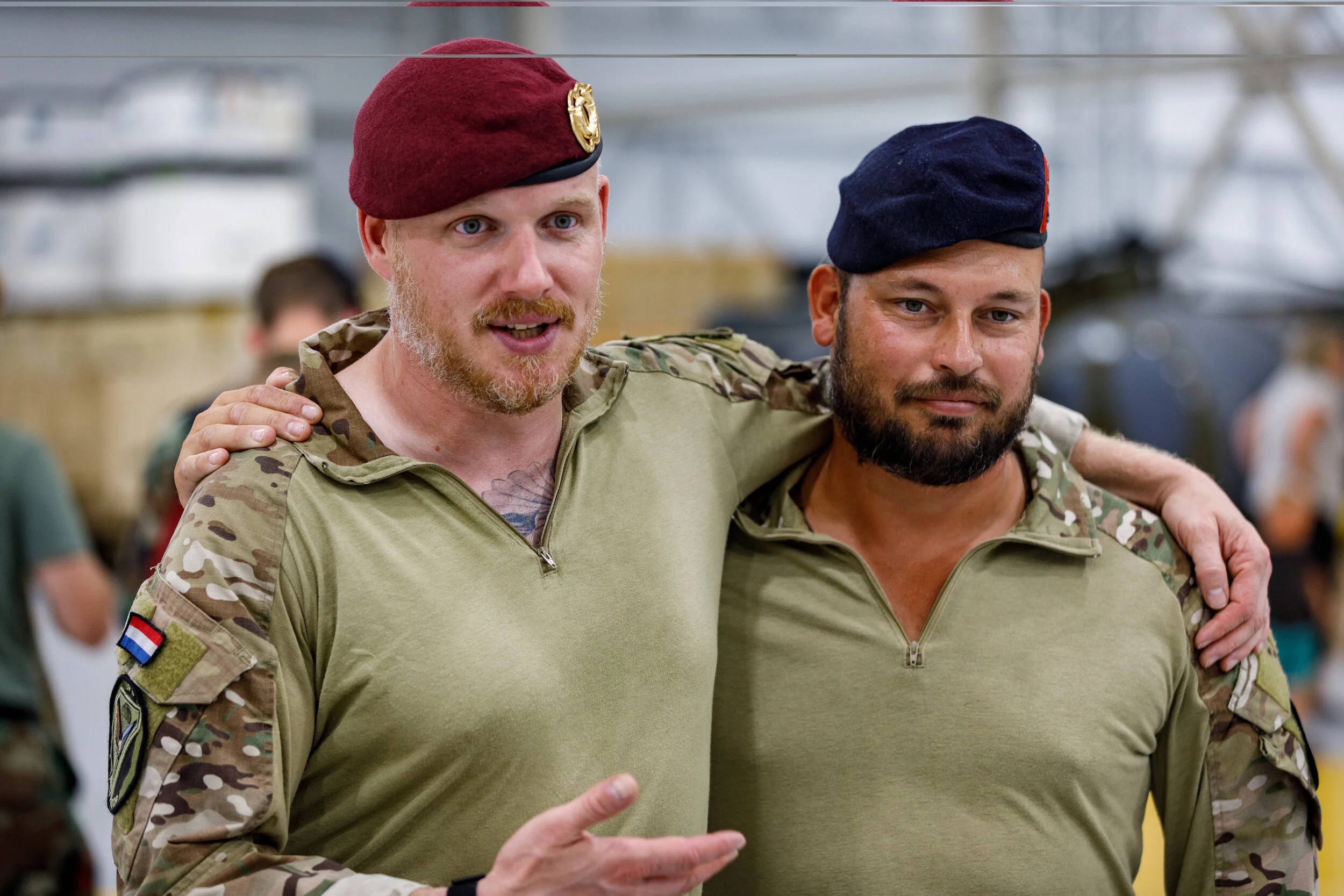 Two men in military uniforms with camouflage patterns and tactical patches, standing indoors with other people in the background. One man is wearing a maroon beret, and the other a navy beret, with the man on the left gesturing with his hand.