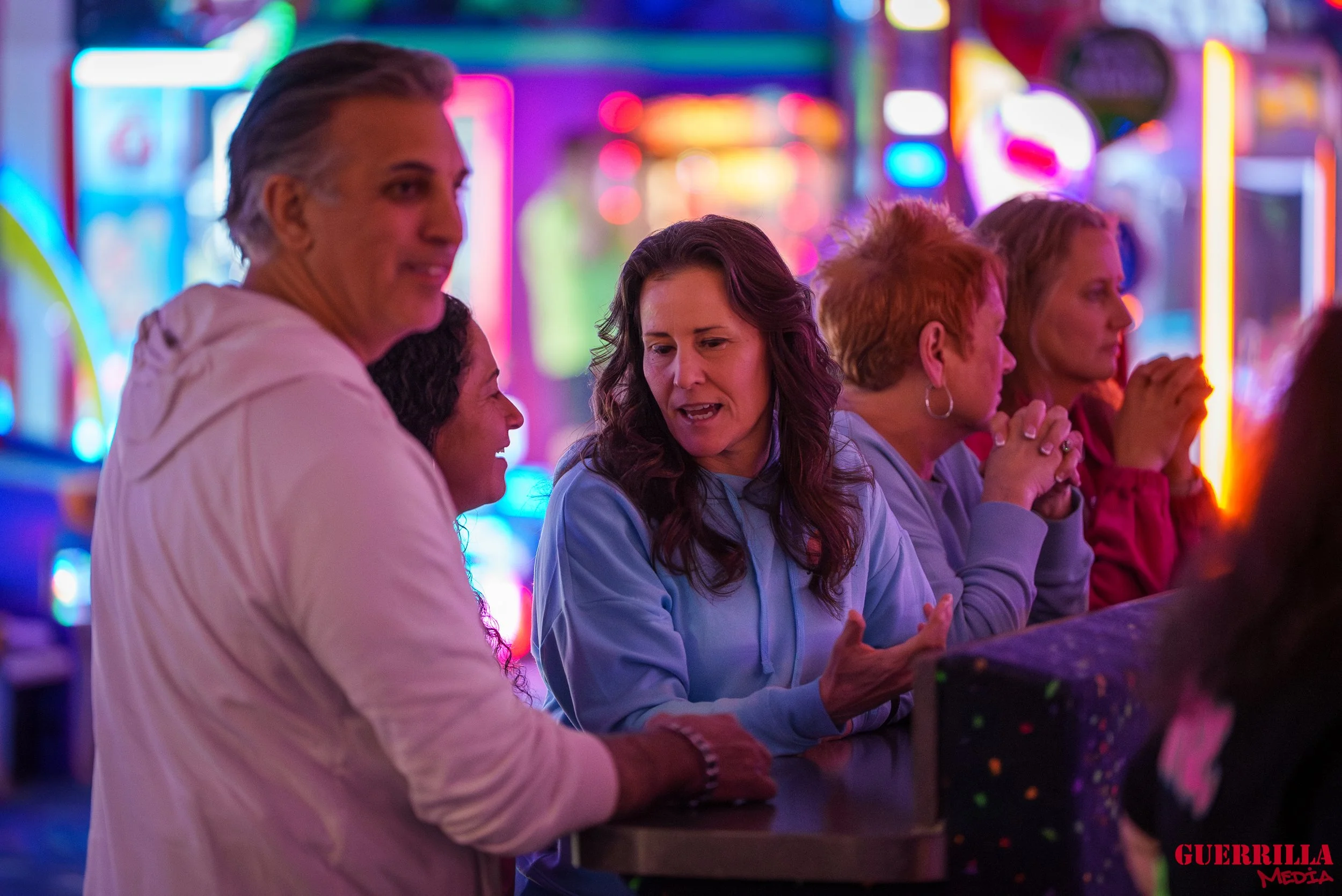 Group of women socializing at an arcade or amusement park with bright, colorful neon lights in the background.
