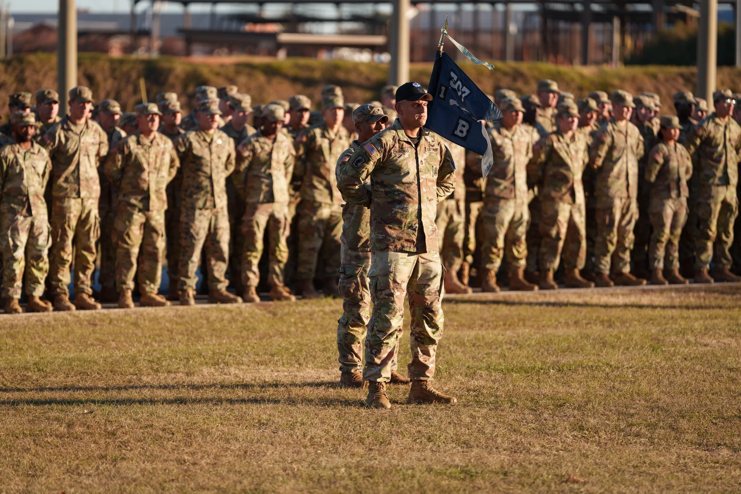 A group of soldiers in camouflage uniforms standing in formation on a field, with one soldier in front holding a flag.