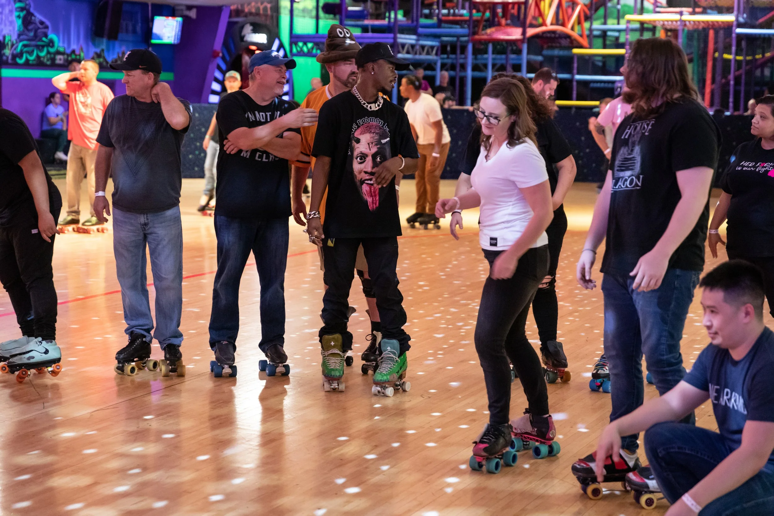 Group of people roller skating indoors at an entertainment center. Some are standing in line, while others are skating or sitting on the floor. The background features colorful railings and a vibrant atmosphere.