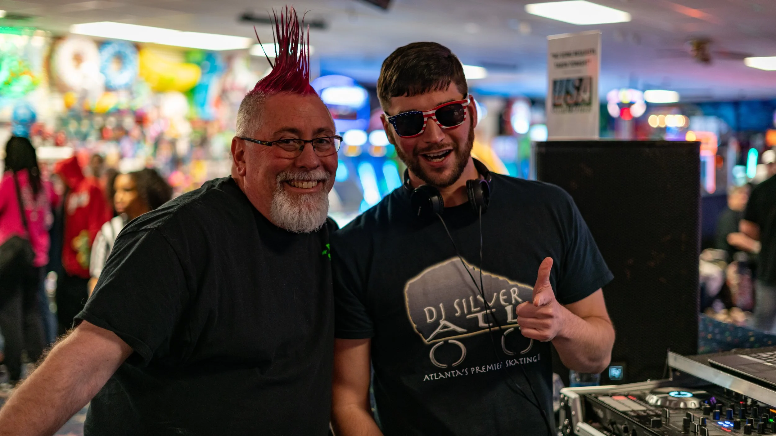 Two men smiling, one with a mohawk hairstyle dyed pink and glasses, and the other wearing sunglasses with an American flag pattern and headphones around his neck, standing in an arcade or gaming center.