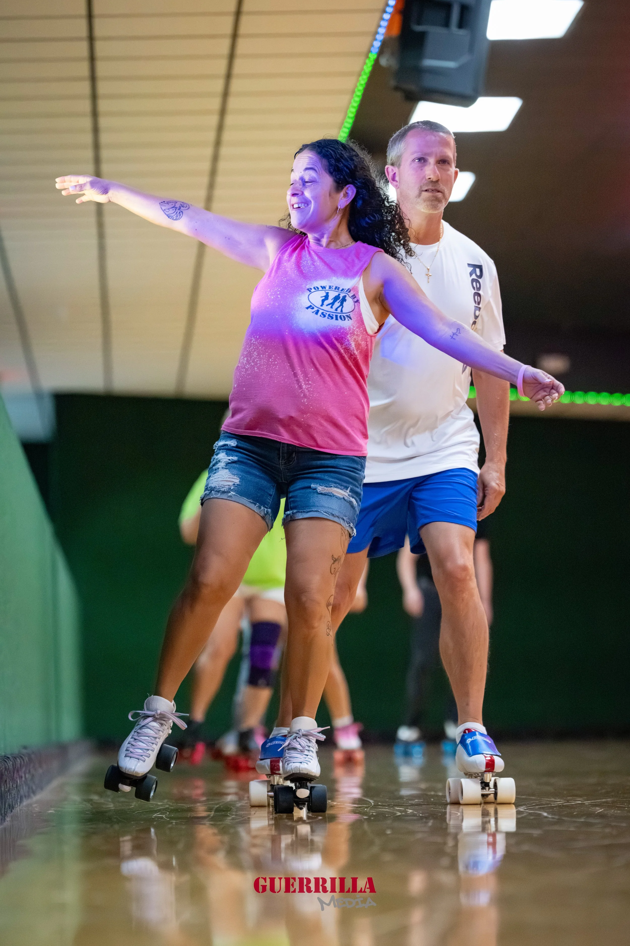 Two people roller skating indoors, a woman in a pink tank top and denim shorts extending her arms, and a man in a white t-shirt and blue shorts behind her. Other skaters are visible in the background.