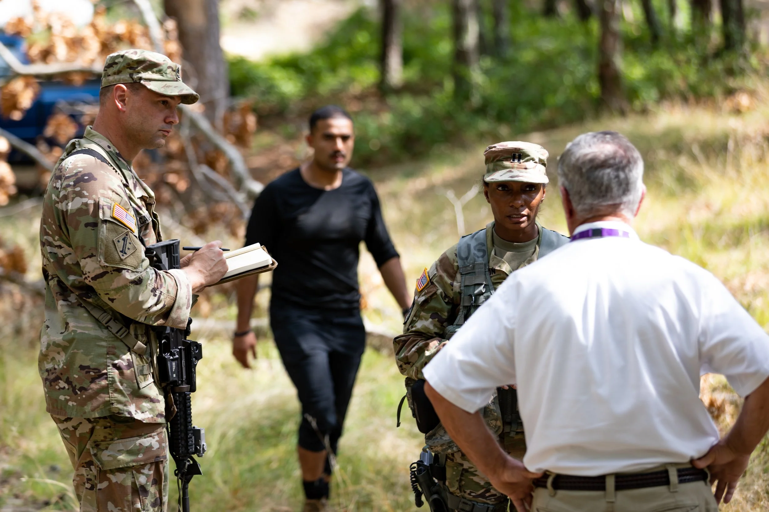 Military personnel and a civilian man having a discussion outdoors in a wooded area.