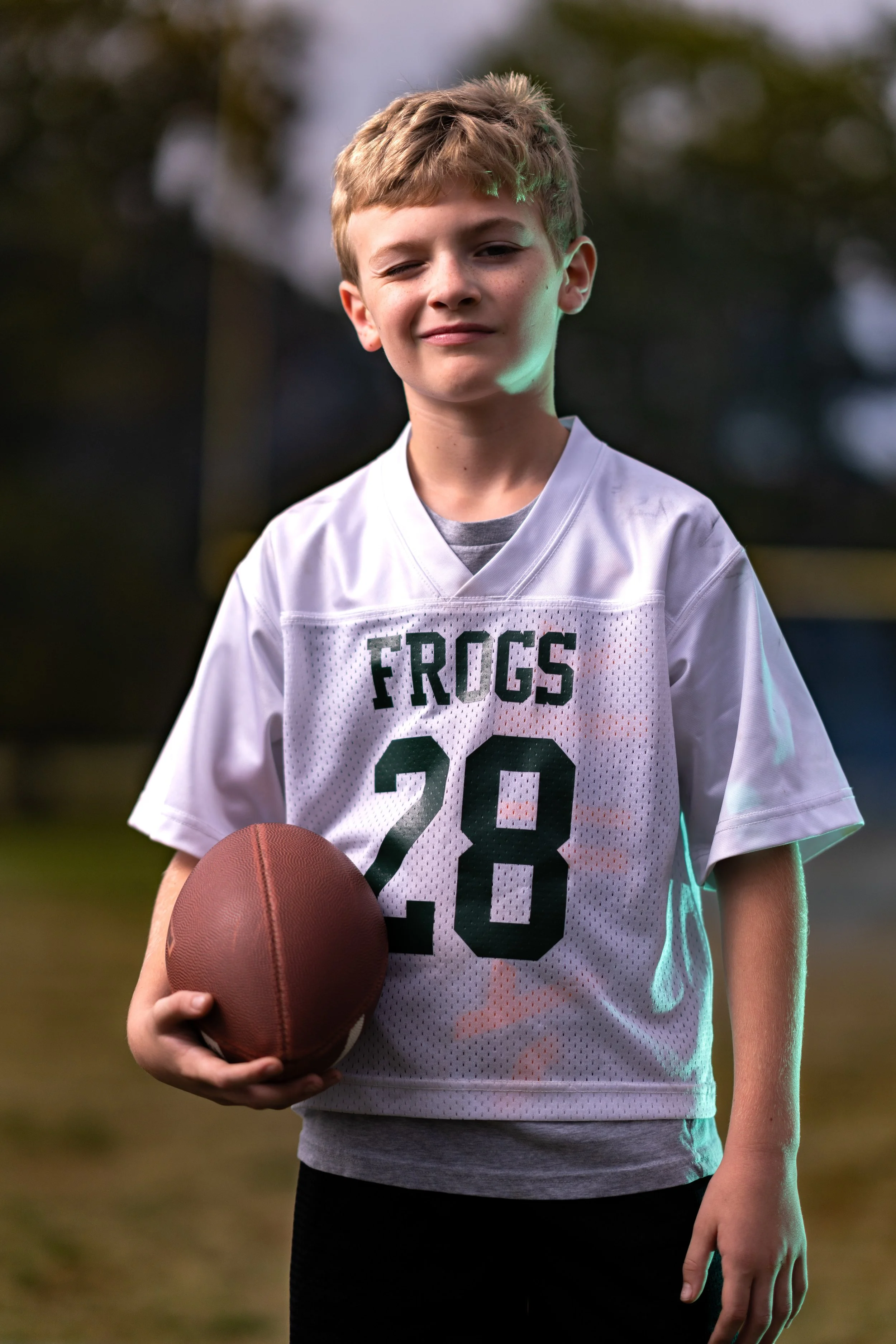 A young boy with short blonde hair in a white football jersey with the number 28 and the word 'FROGS' on it, holding a football, standing outdoors on a grassy field.
