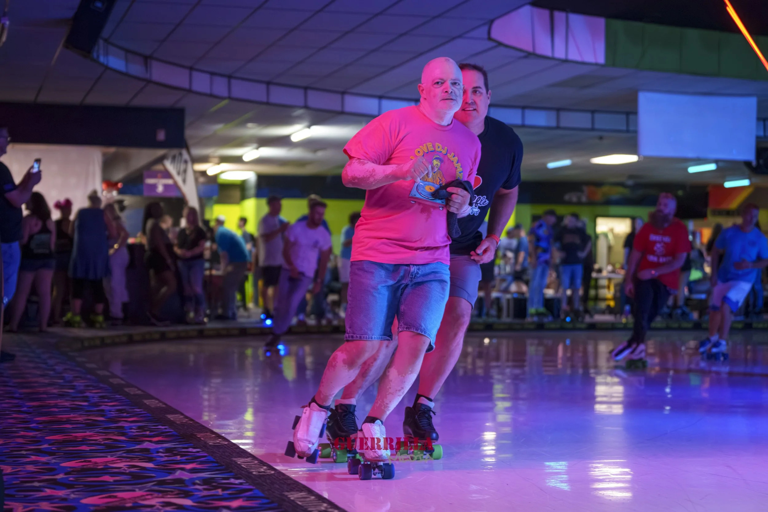 Two men roller skating in a colorful indoor skating rink with a crowd of people in the background. One man is wearing a pink T-shirt and denim shorts, and the other is in a black T-shirt and shorts, both smiling and enjoying themselves.