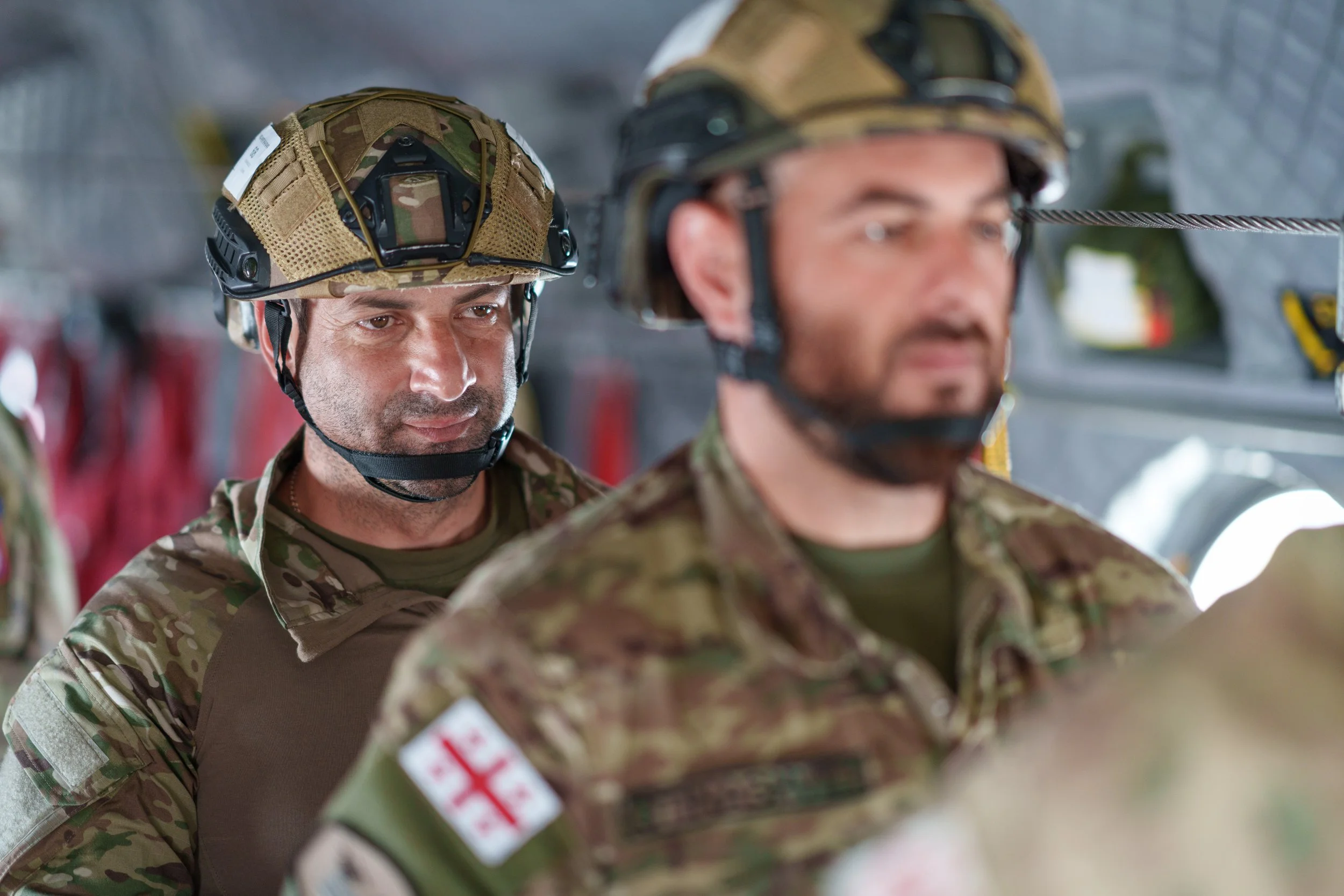 Two soldiers in camouflage uniforms and helmets standing in a military setting, with a third soldier partially visible in the foreground.