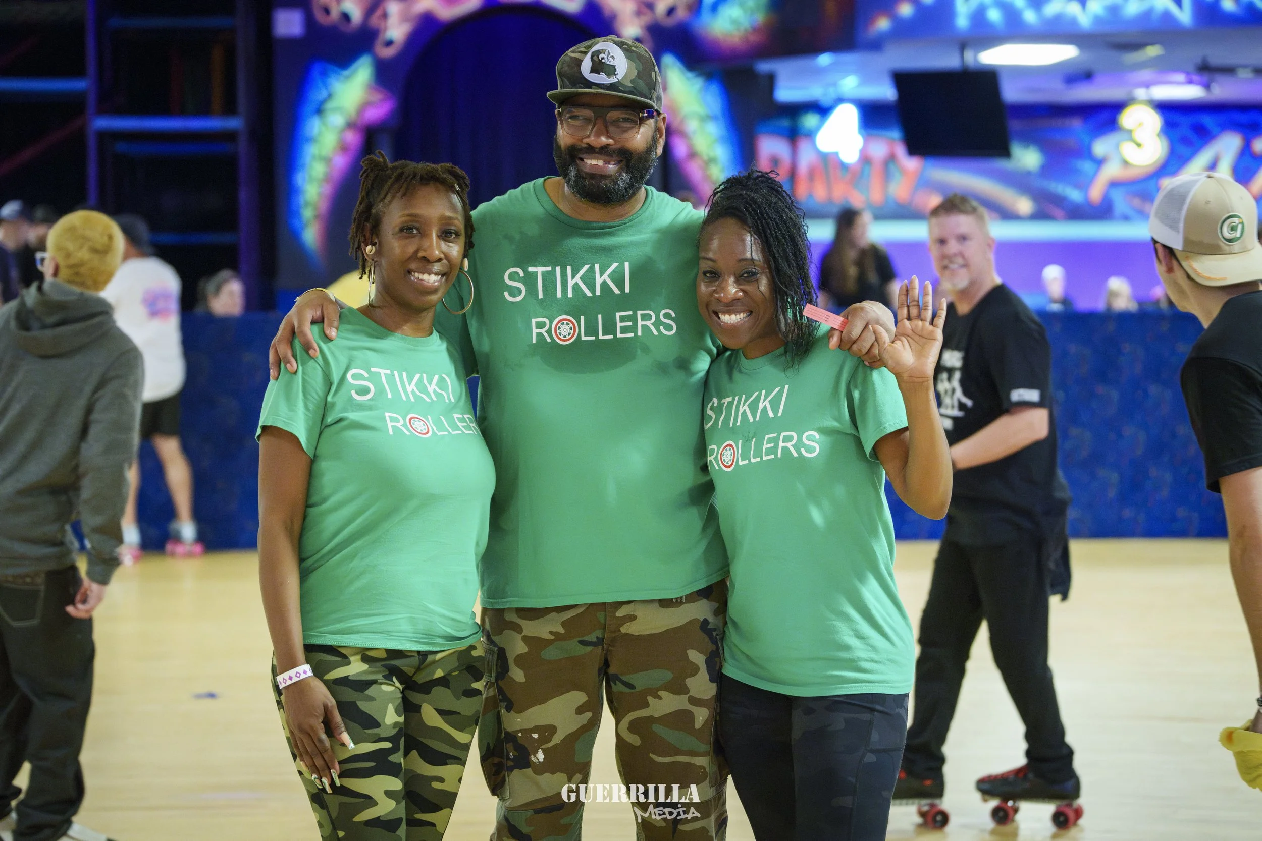 Three smiling people in green T-shirts that read 'STIKKI ROLLERS', posing together at roller skating rink, with other skaters and colorful lights in the background.