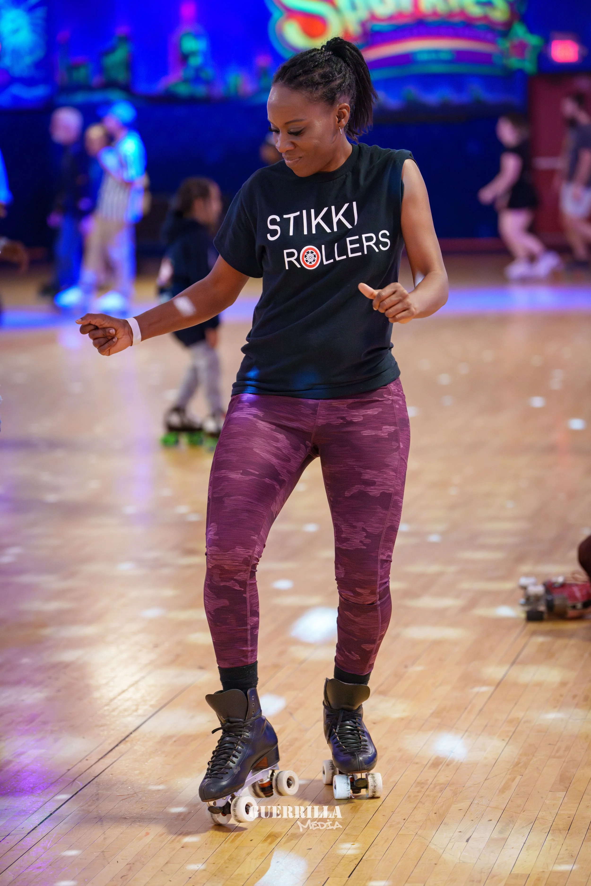 Woman roller skating indoors on a wooden floor, wearing a black T-shirt that says 'STIKKI ROLLERS', purple camouflage leggings, and black roller skates. She appears to be dancing or practicing at a roller skating rink.