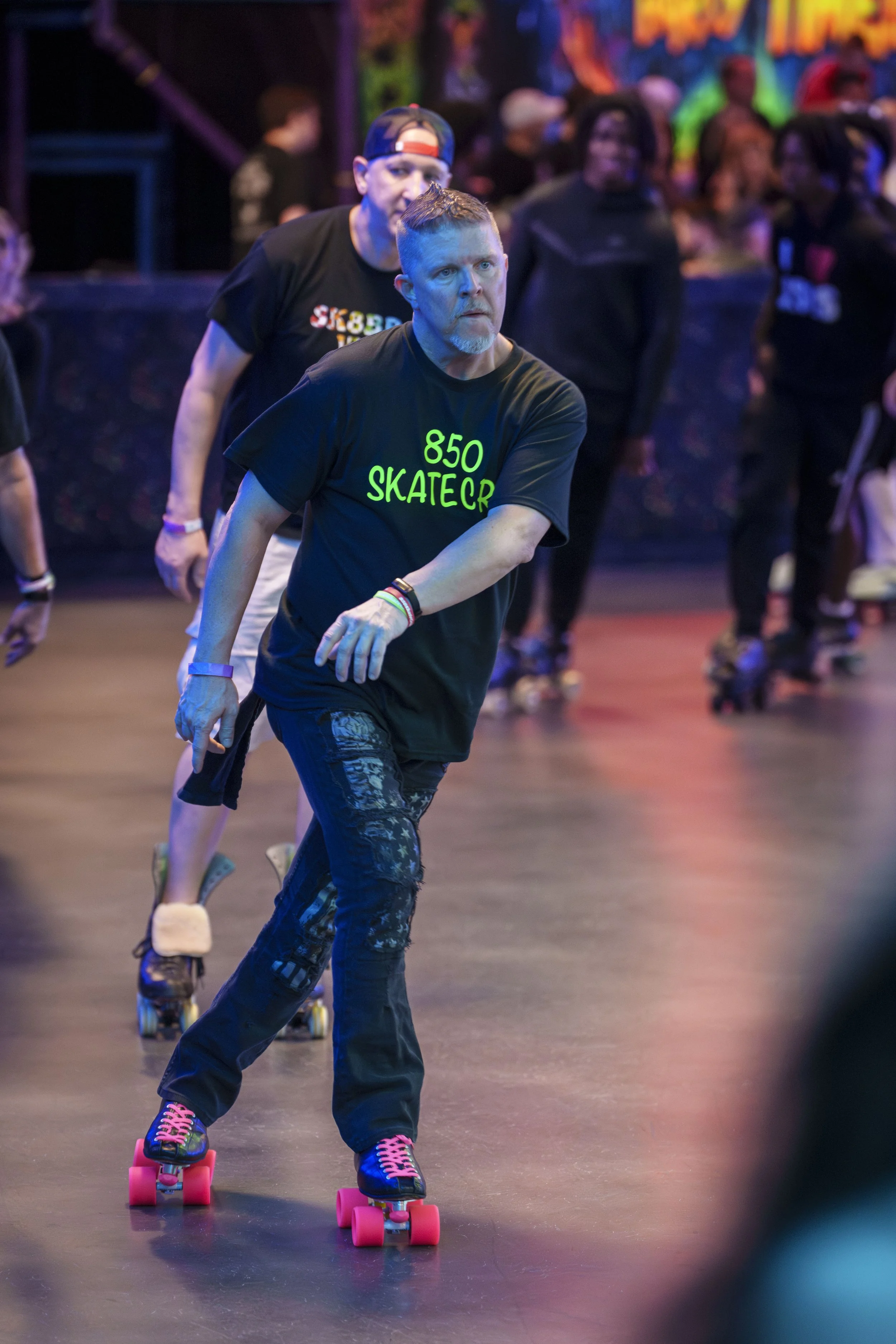 A man roller skating at an indoor roller rink, wearing a black T-shirt that says '850 SKATER' in green letters, with a colorful background and other skaters in the background.