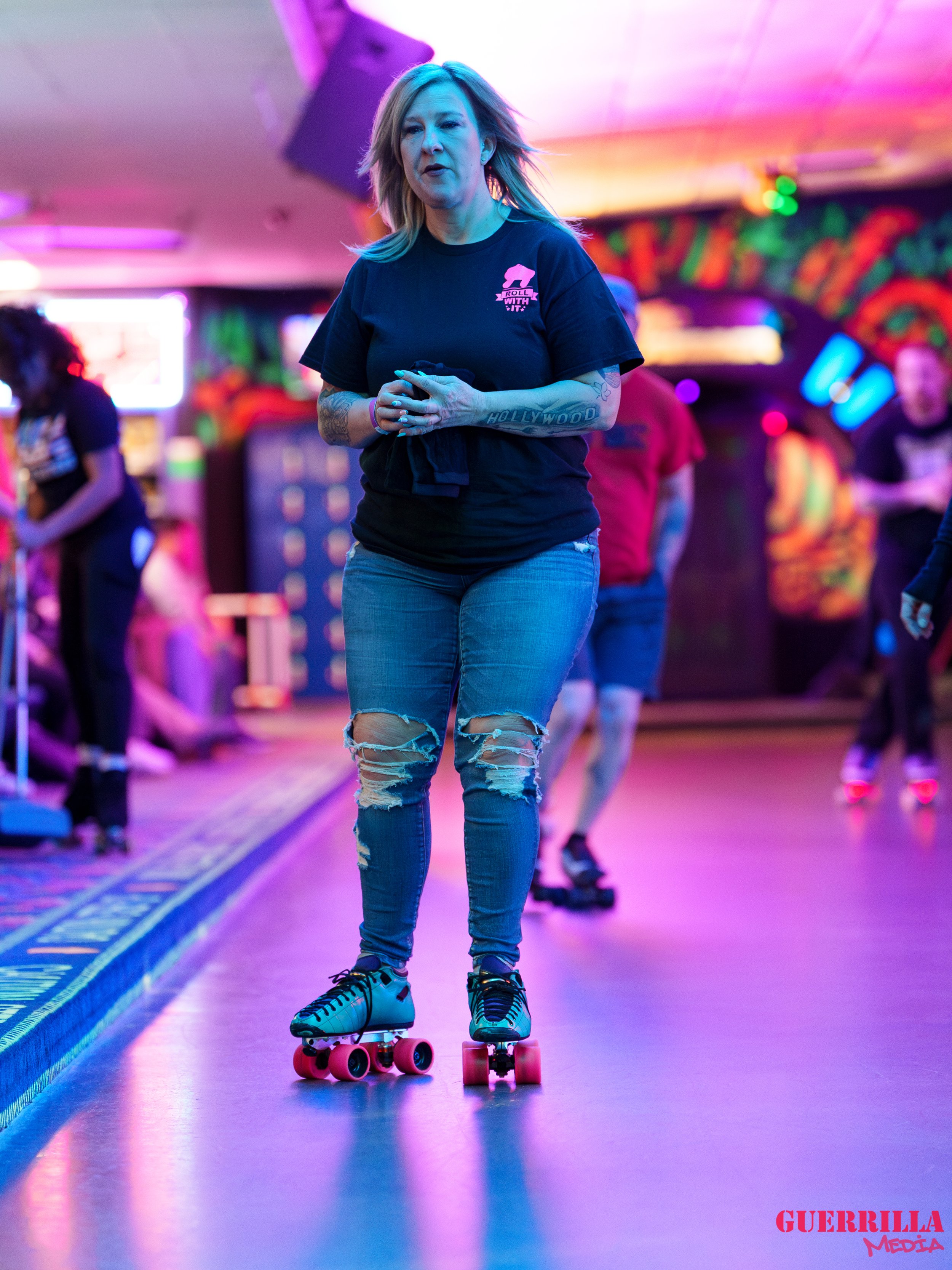 A woman roller skating in an indoor roller rink with colorful neon lighting, wearing ripped jeans and a black t-shirt.