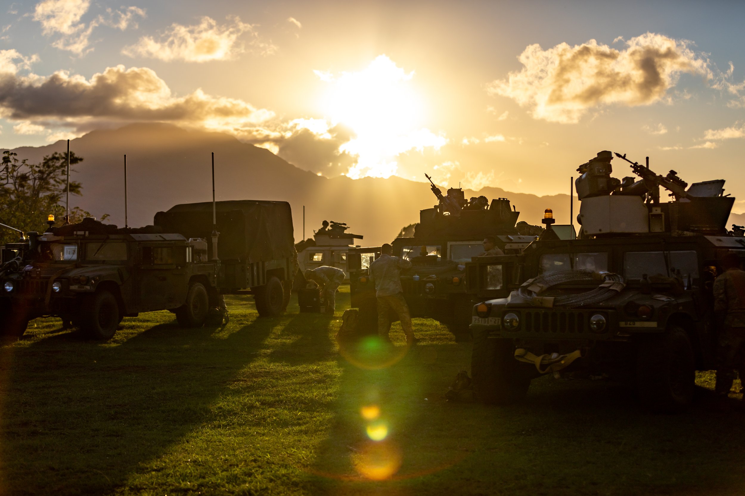 Group of military vehicles and soldiers during sunset on grassy field with mountains in background.