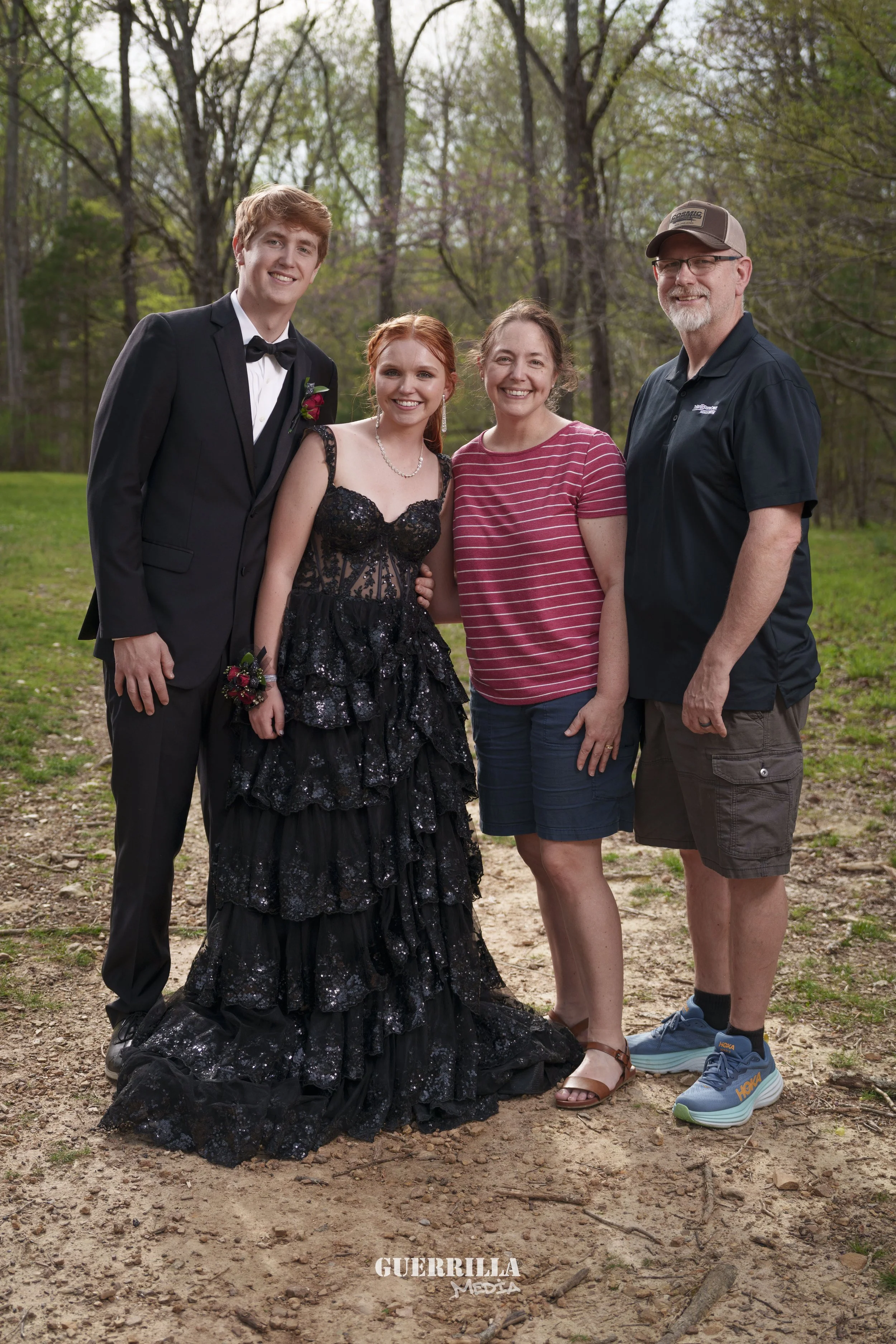 A family of four standing outdoors in a park during daytime, with trees and grass in the background. The son is dressed in a black tuxedo with a bow tie, the daughter is wearing a black, lace, ruffled gown, the mother is in a red and white striped sh