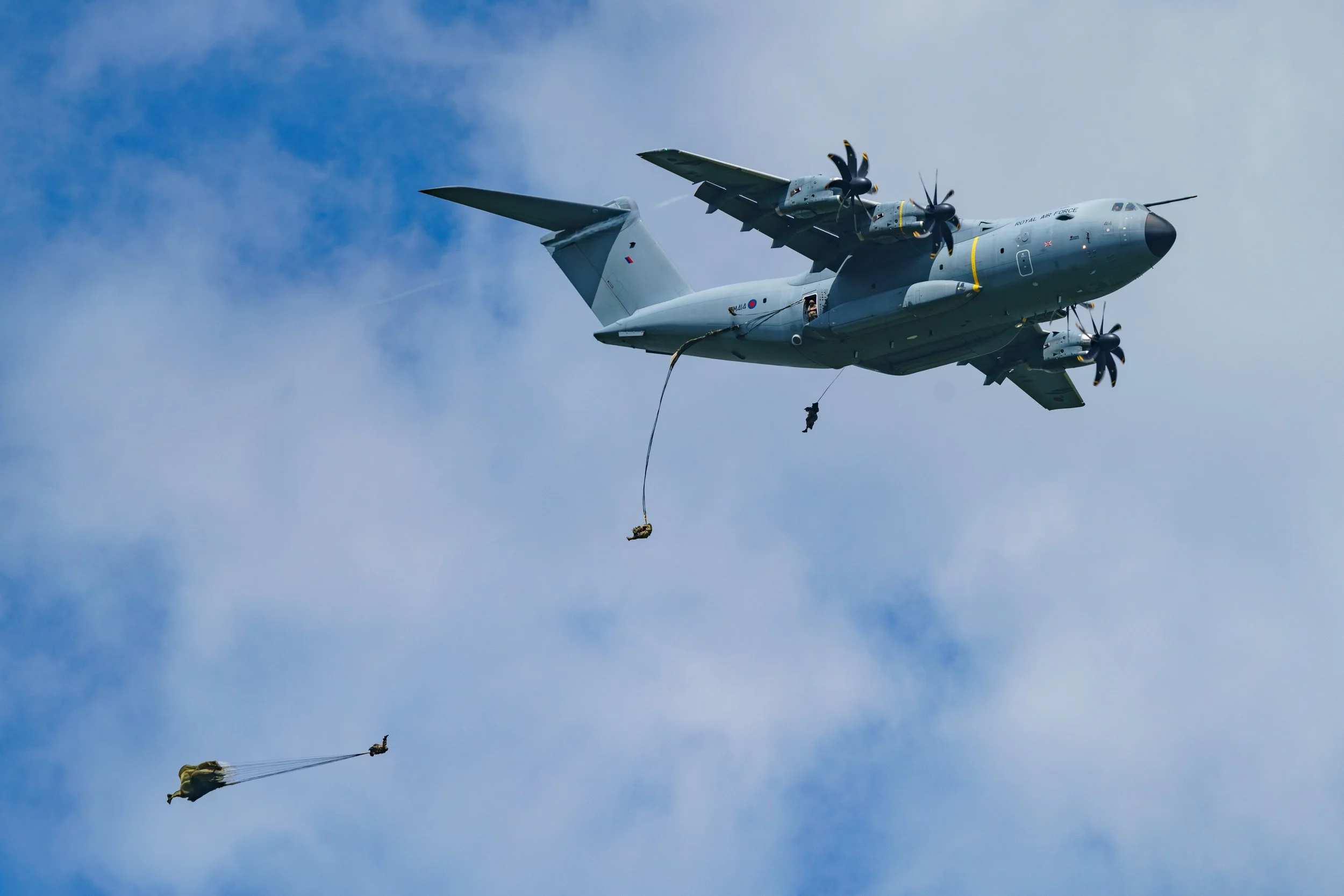 A military aircraft, possibly a type of cargo plane, in flight with skydivers exiting the aircraft.