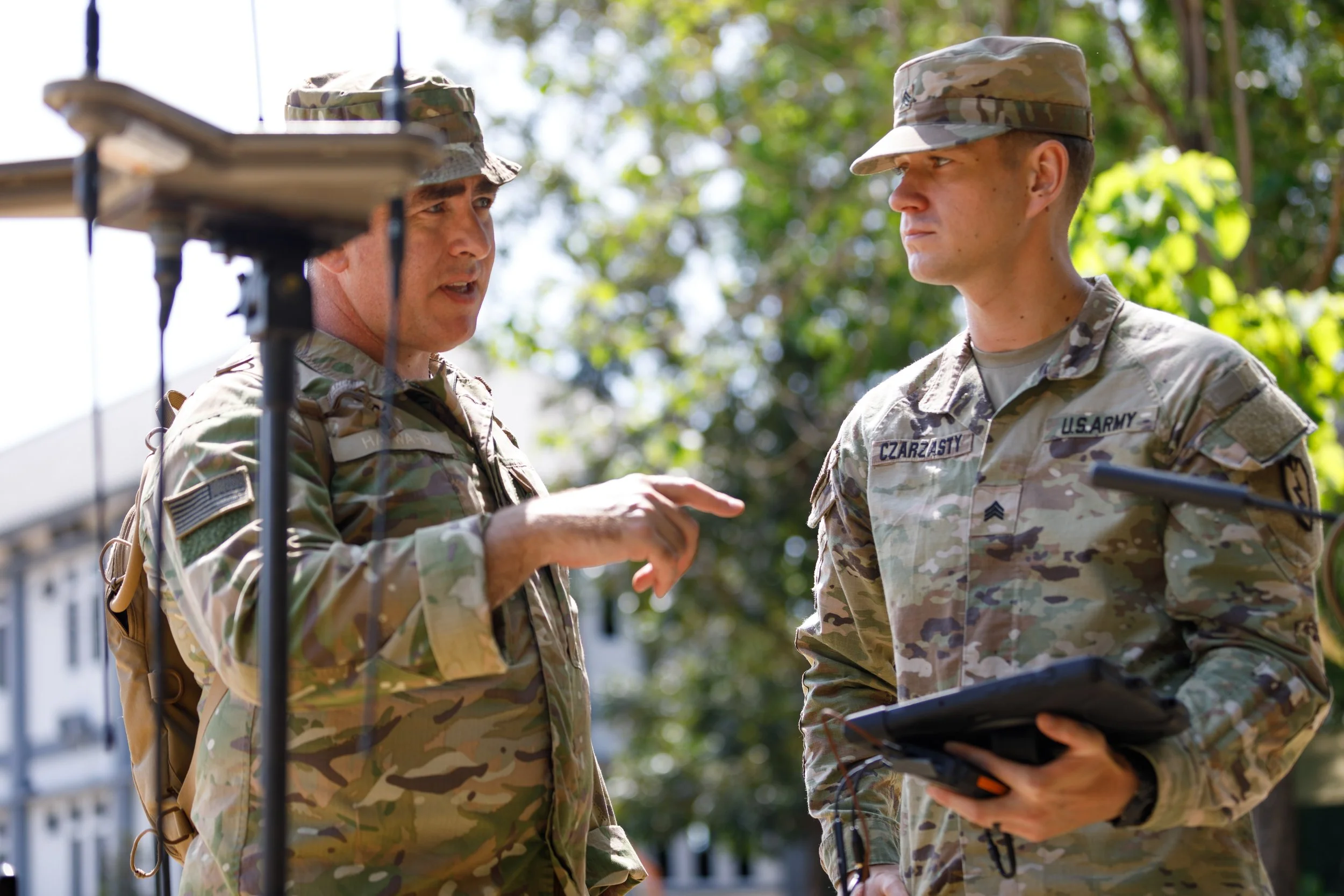 Two soldiers in camouflage uniforms having a discussion outdoors, with one pointing at a tablet device, surrounded by trees and a building.
