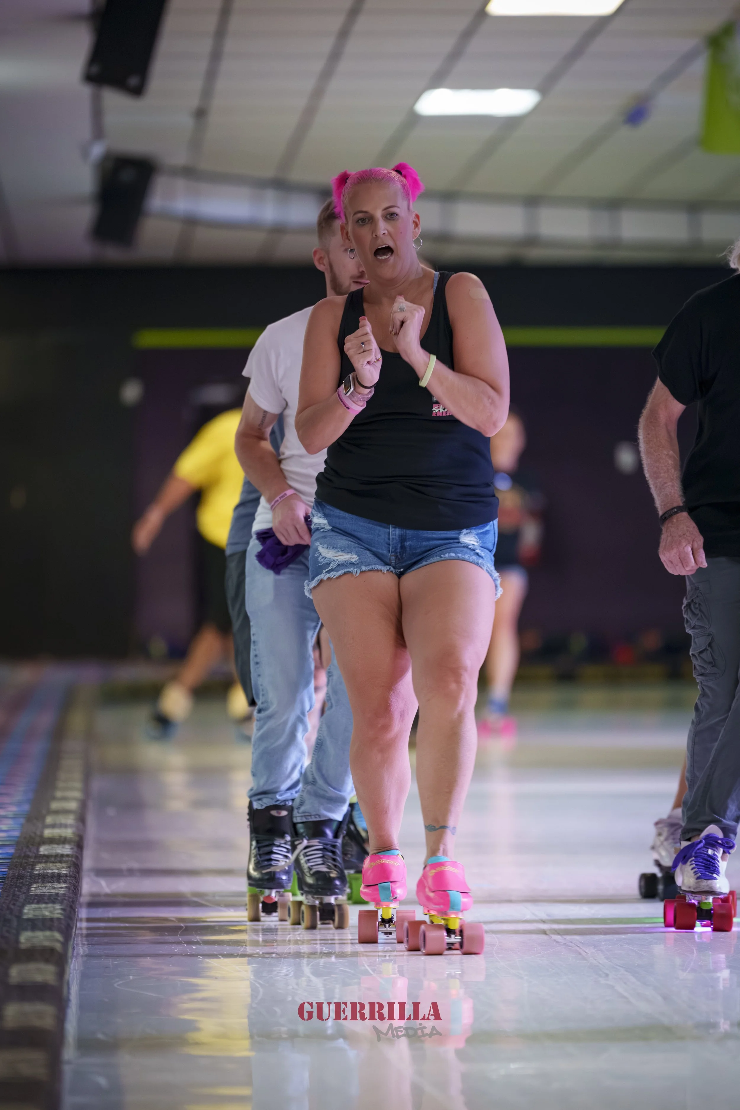 Woman with pink pigtails roller skating indoors, wearing a black tank top and denim shorts, surrounded by other skaters.