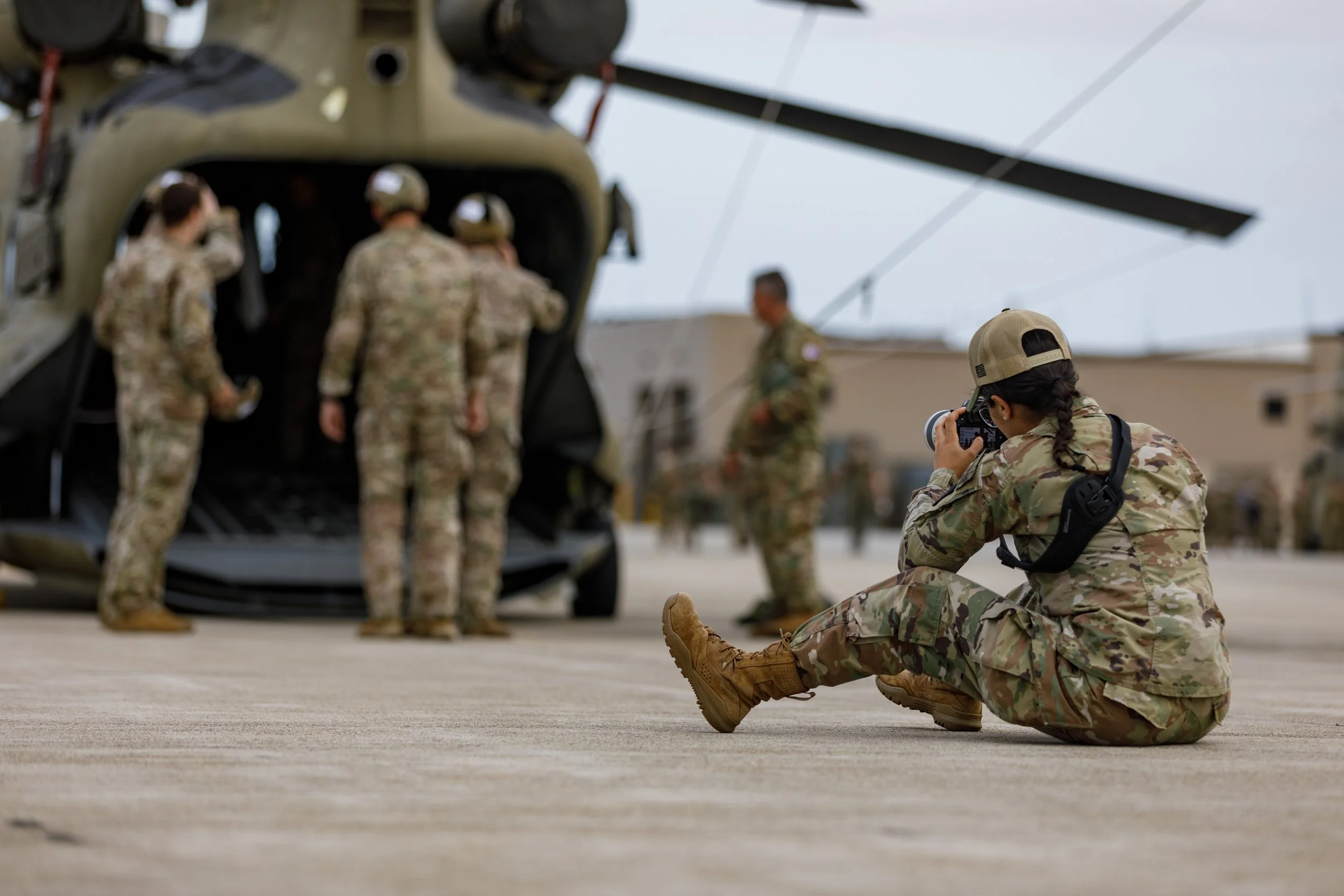 A female soldier in camouflage uniform sitting on the ground taking a photo of a group of soldiers in camouflage uniform near a military helicopter on an airstrip.