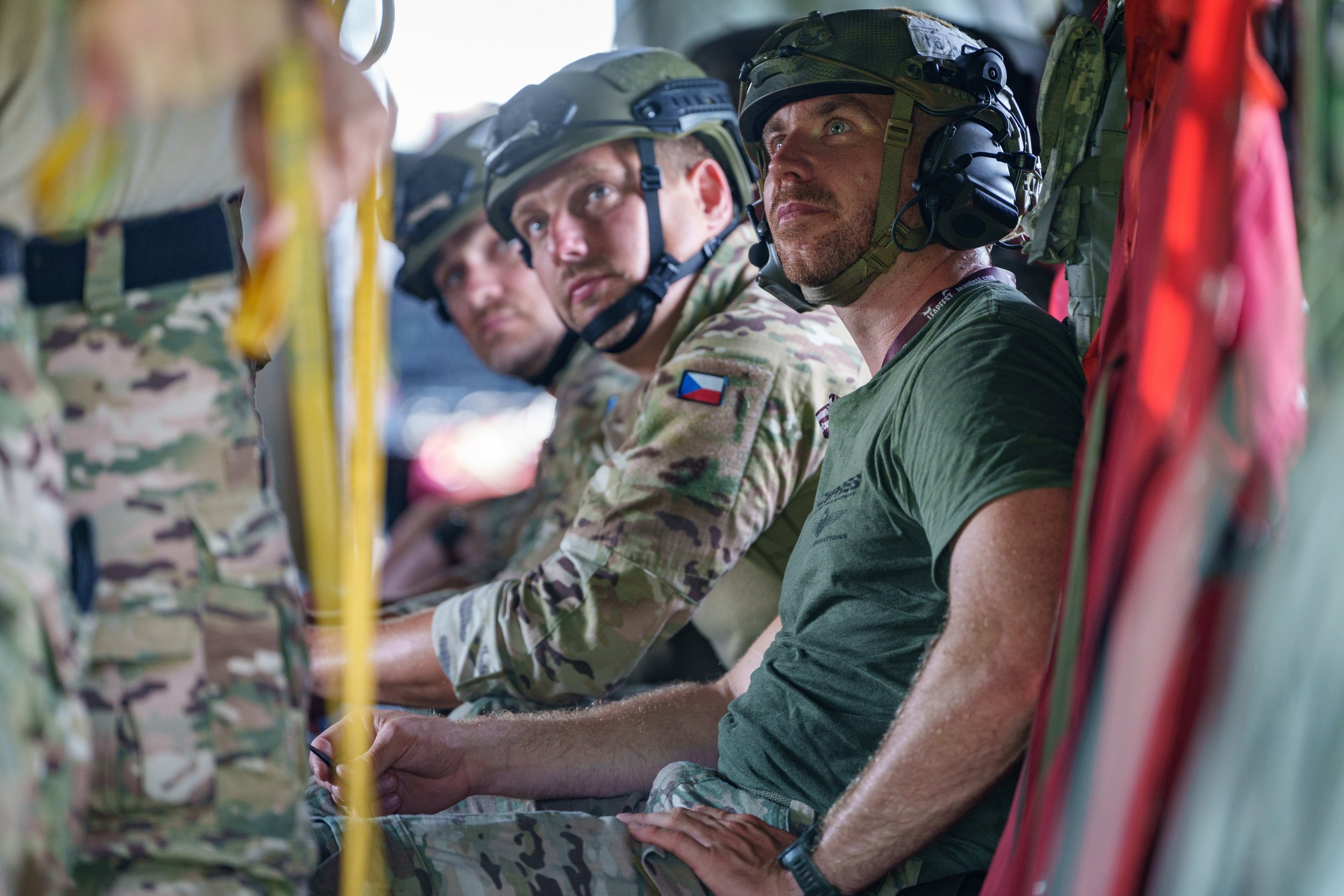 Military soldiers sitting in a helicopter wearing helmets and camouflage uniforms, with one soldier in a green t-shirt.