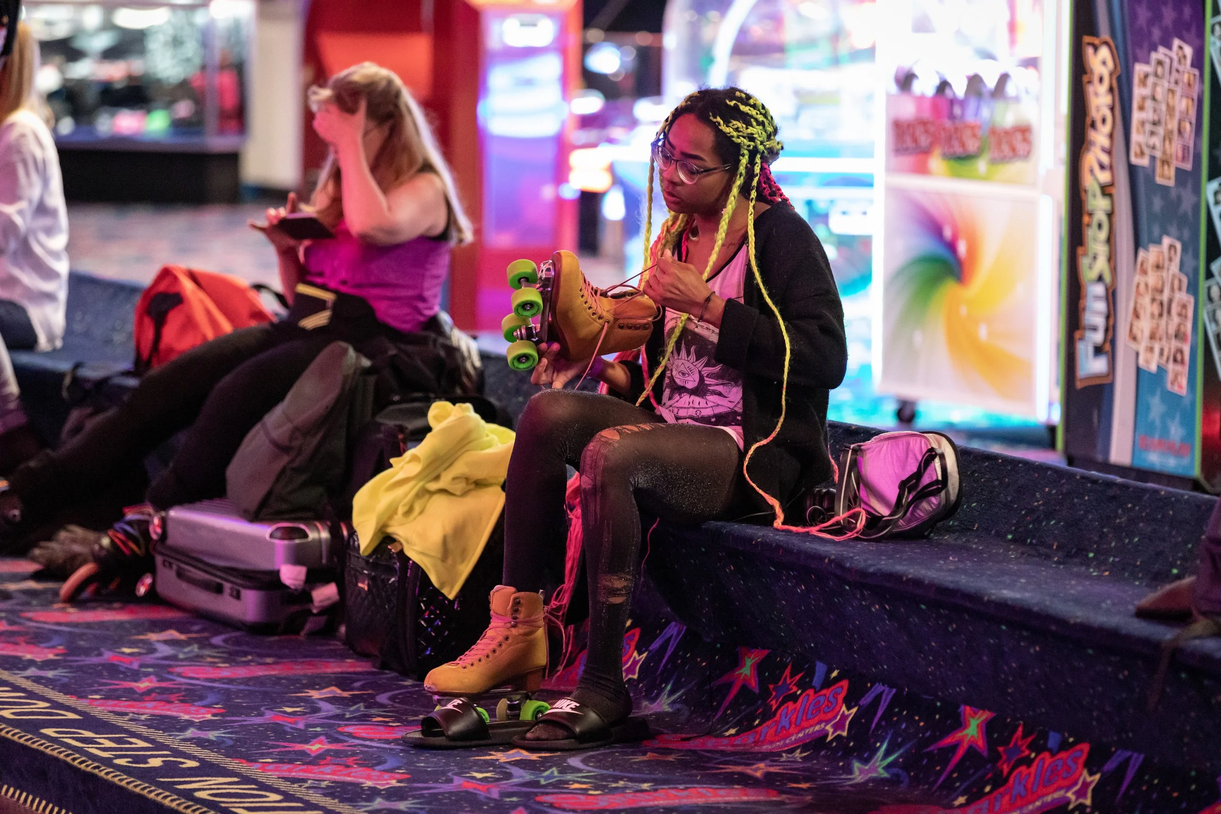 A young woman with yellow and pink braided hair sitting on a bench at an arcade, tying her roller skate. She is wearing glasses, a black cardigan, and black leggings. She has a pink and white shirt with a graphic design, and yellow roller skates. The