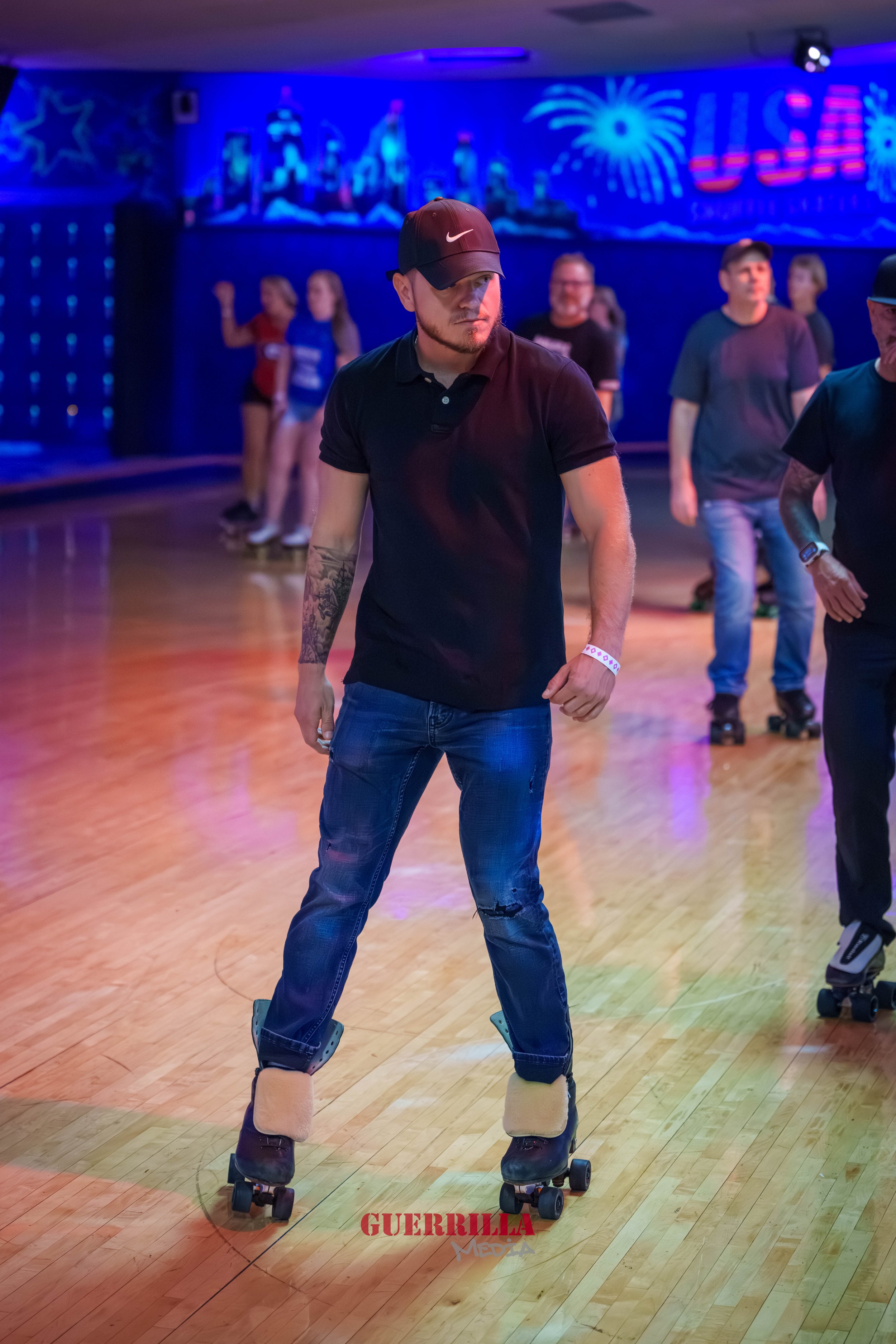 A man roller skating in a roller rink with a wooden floor and colorful neon lights in the background. He is wearing a black polo shirt, blue jeans with a rip on the knee, a dark cap, and wristbands.