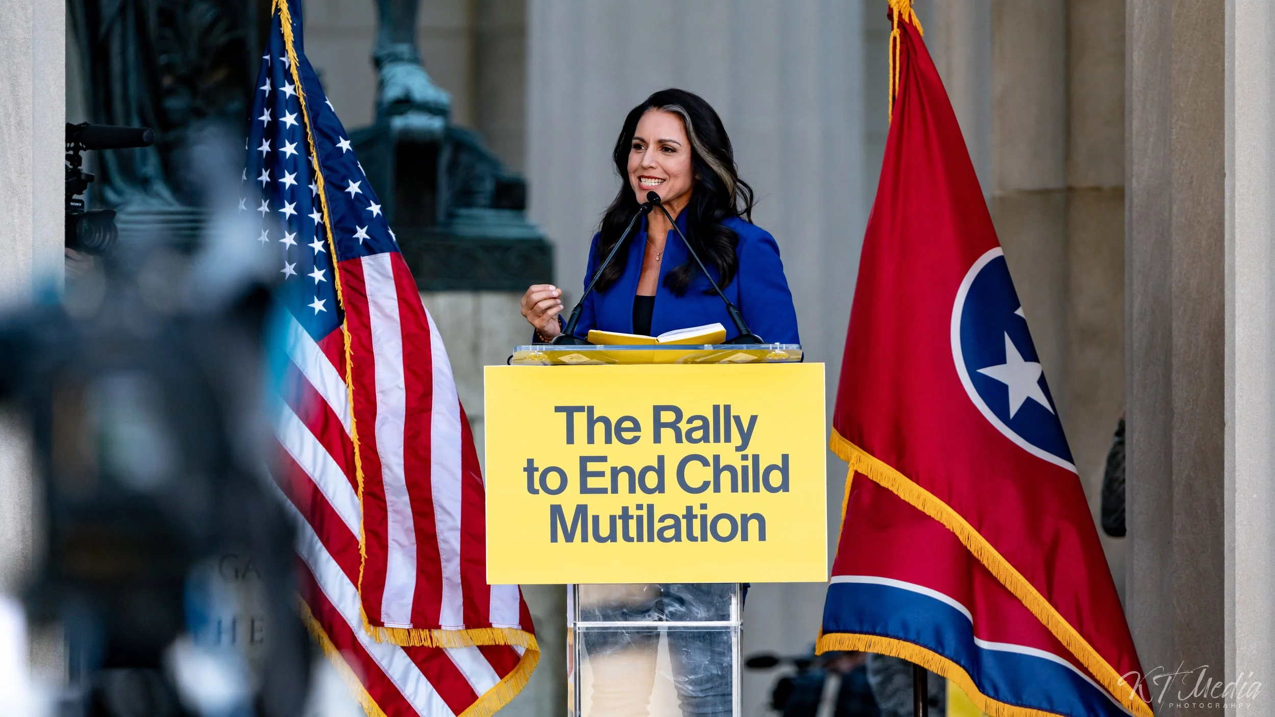 A woman in a blue blazer speaking at a podium during a rally for ending child mutilation, with American and Tennessee flags behind her.
