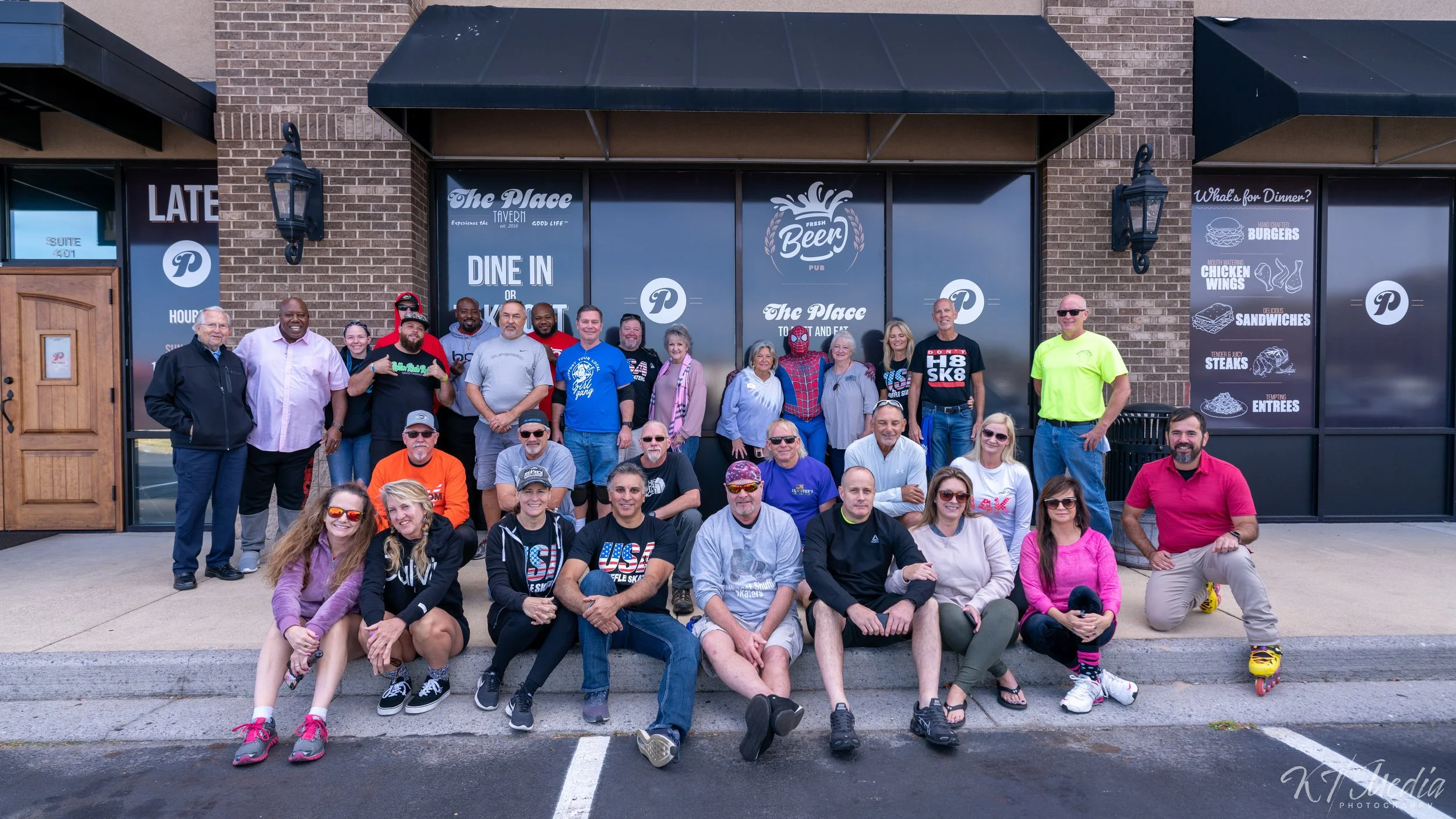 Group of people gathered outside a restaurant called 'The Place', some sitting on the curb and others standing, with signage advertising menu items like burgers, chicken wings, sandwiches, steaks, and entrees, in front of a brick building with black 