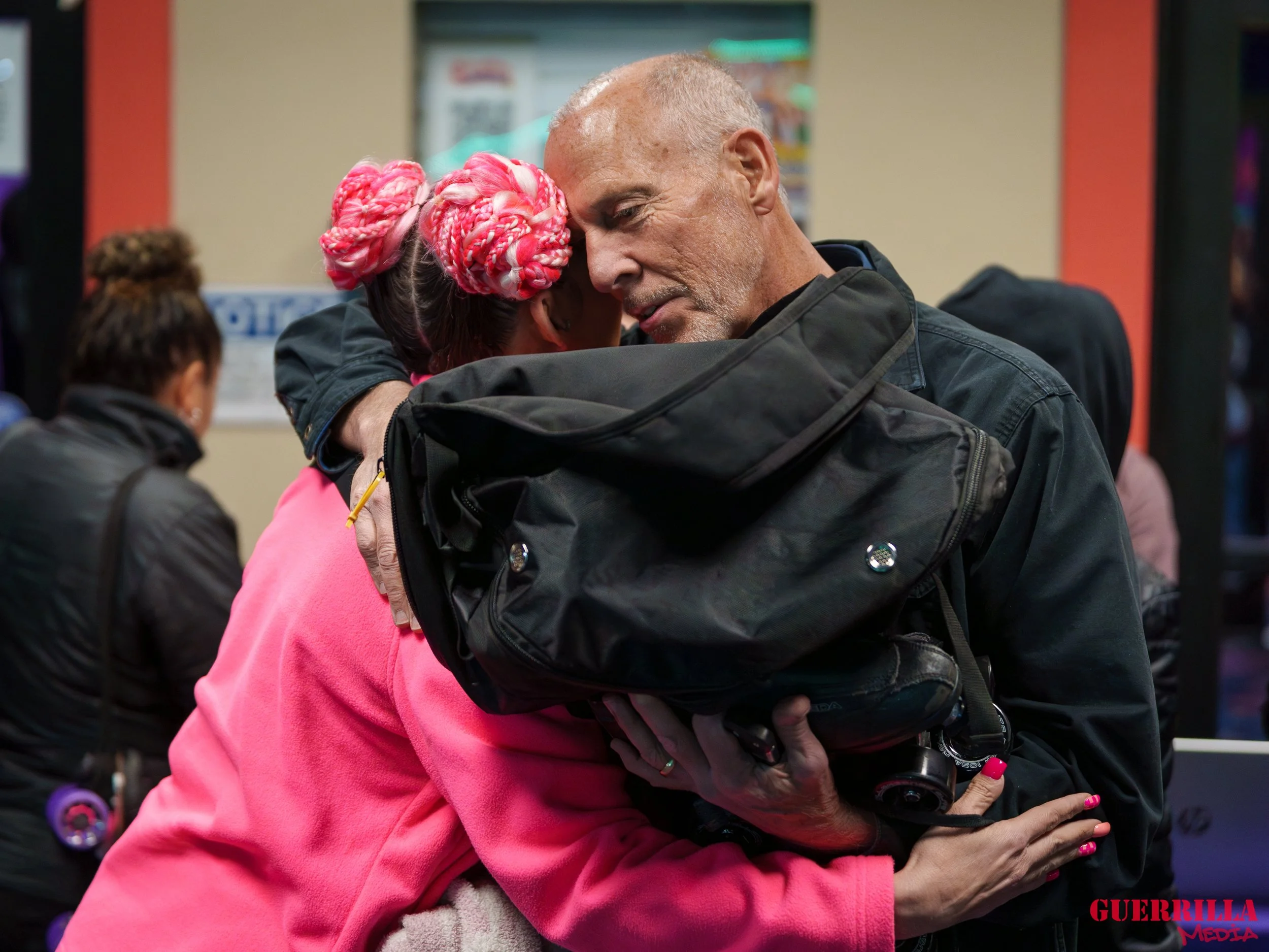 An older man and a woman sharing a heartfelt hug in a public place, with the woman wearing pink with two pink braided hairpieces.