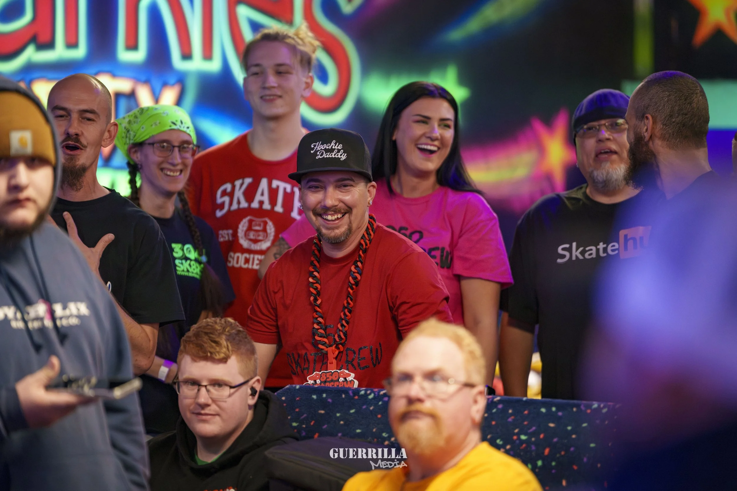 Group of people smiling and enjoying themselves at an indoor skateboarding event with colorful neon lights in the background.