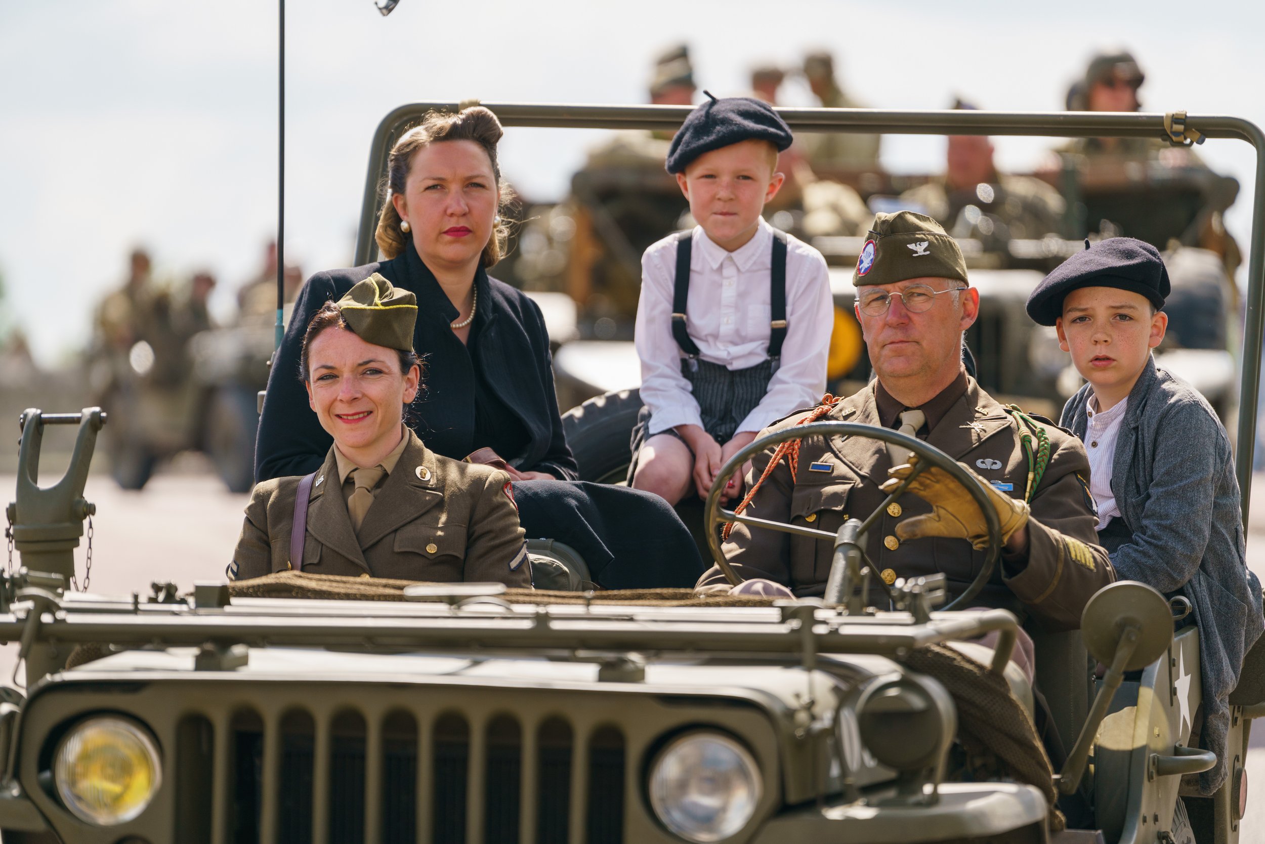 Group of people, including two women and three children, dressed in vintage military and civilian clothing, sitting in a World War II-style military vehicle with a backdrop of soldiers and vehicles during a parade or historical reenactment.