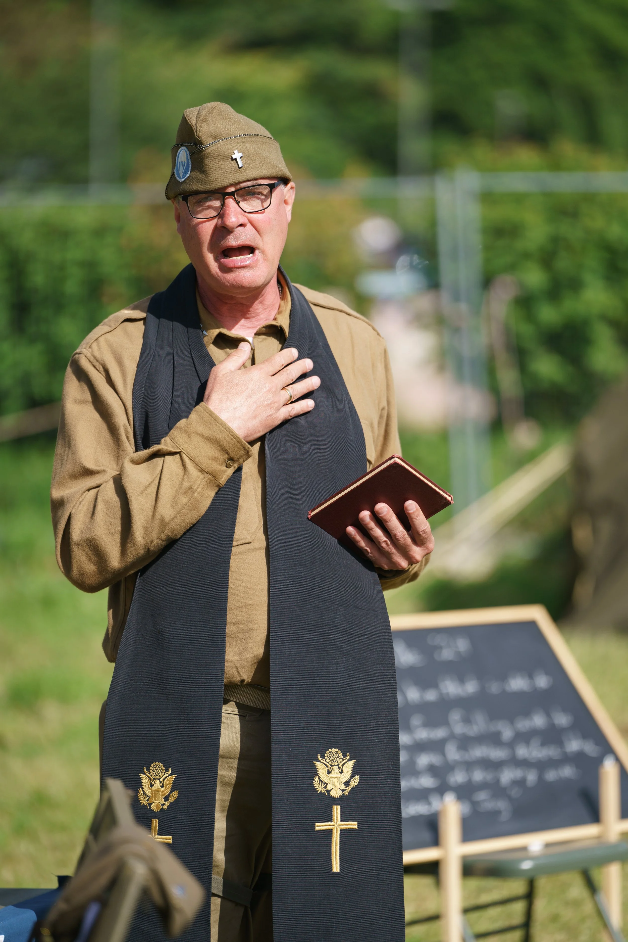 A man in WWII uniform with glasses, a brown hat with patches, a tan shirt, and a black stole with gold religious symbols, standing outdoors with his hand on his chest, holding a prayer book. A blackboard with writing is in the background.