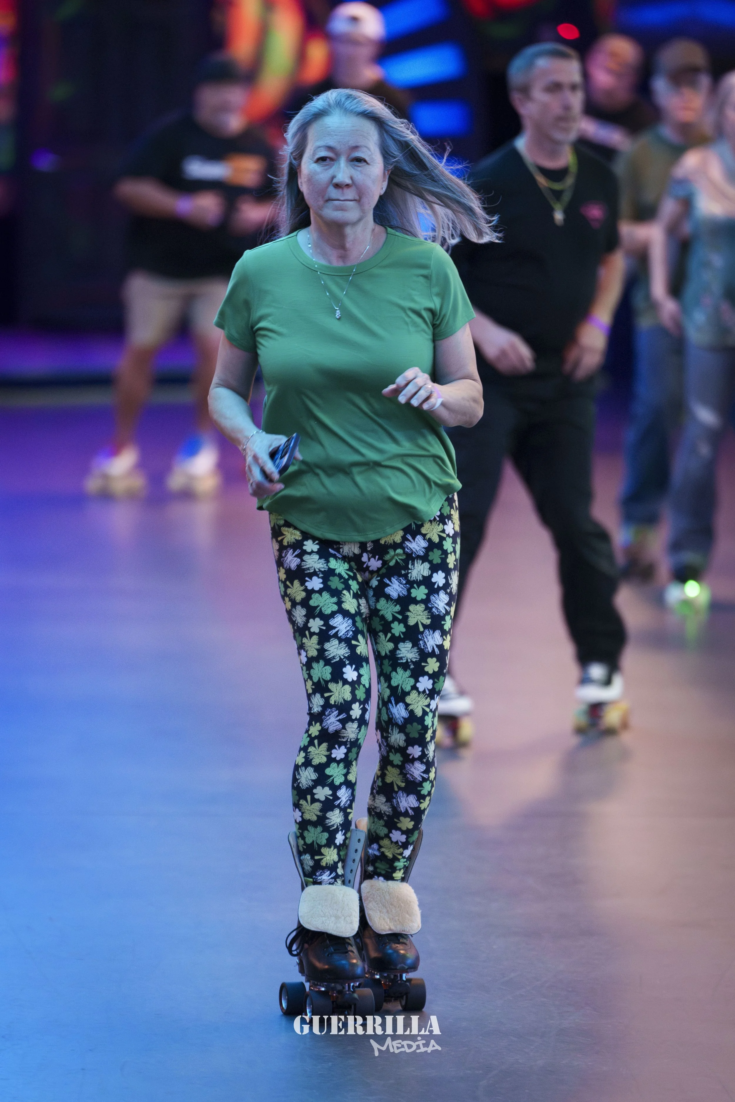 An older woman roller skating at an indoor roller rink, wearing a green t-shirt and patterned leggings with shamrocks.
